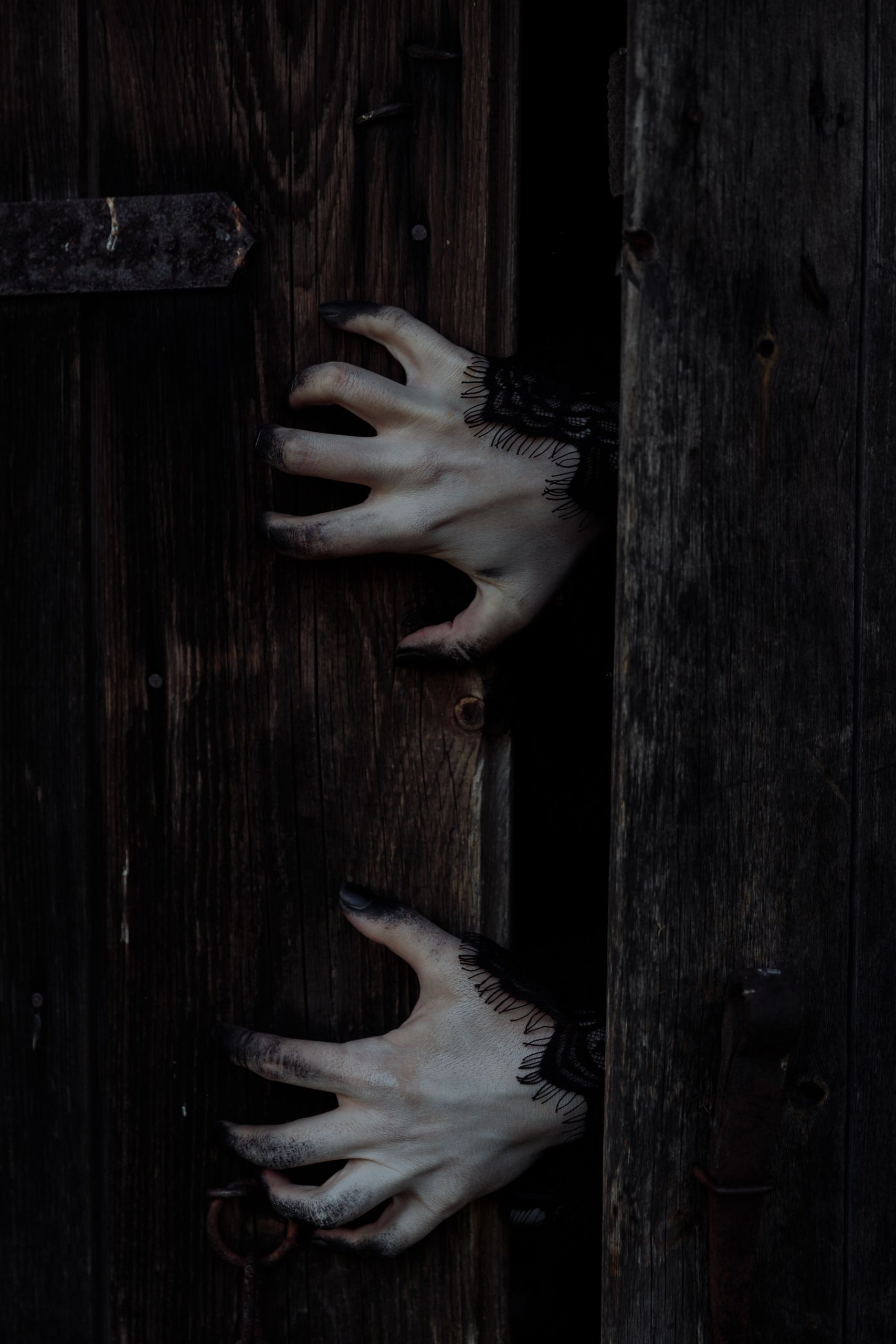 A pair of hands with black fingers and nails are reaching through a wooden door. - Creepy