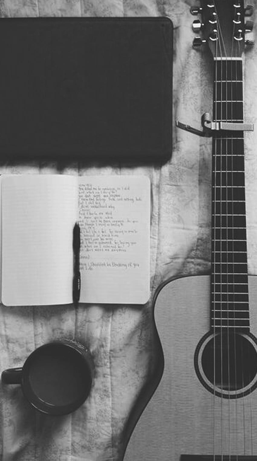 An open notebook with lyrics written in pen, a pen, a cup of coffee, and a guitar. - Guitar
