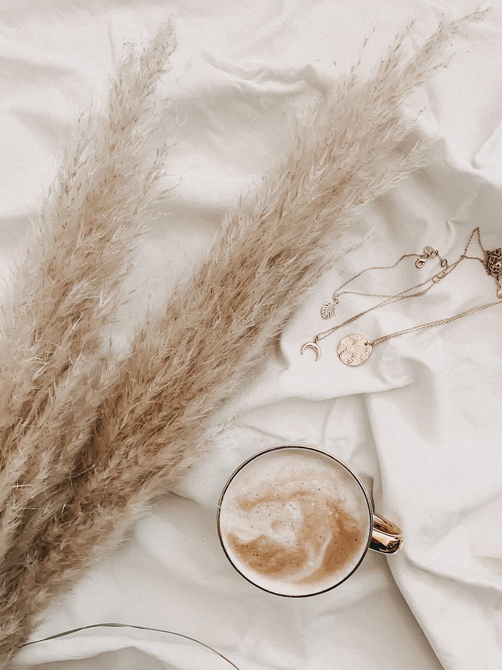 A flat lay of a cup of coffee, pampas grass, and gold necklaces on a white sheet. - Neutral, beige, light brown, cream, brown