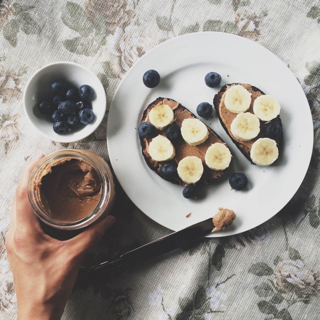 A person holding a jar of peanut butter next to a plate with toast and fruit. - Foodie