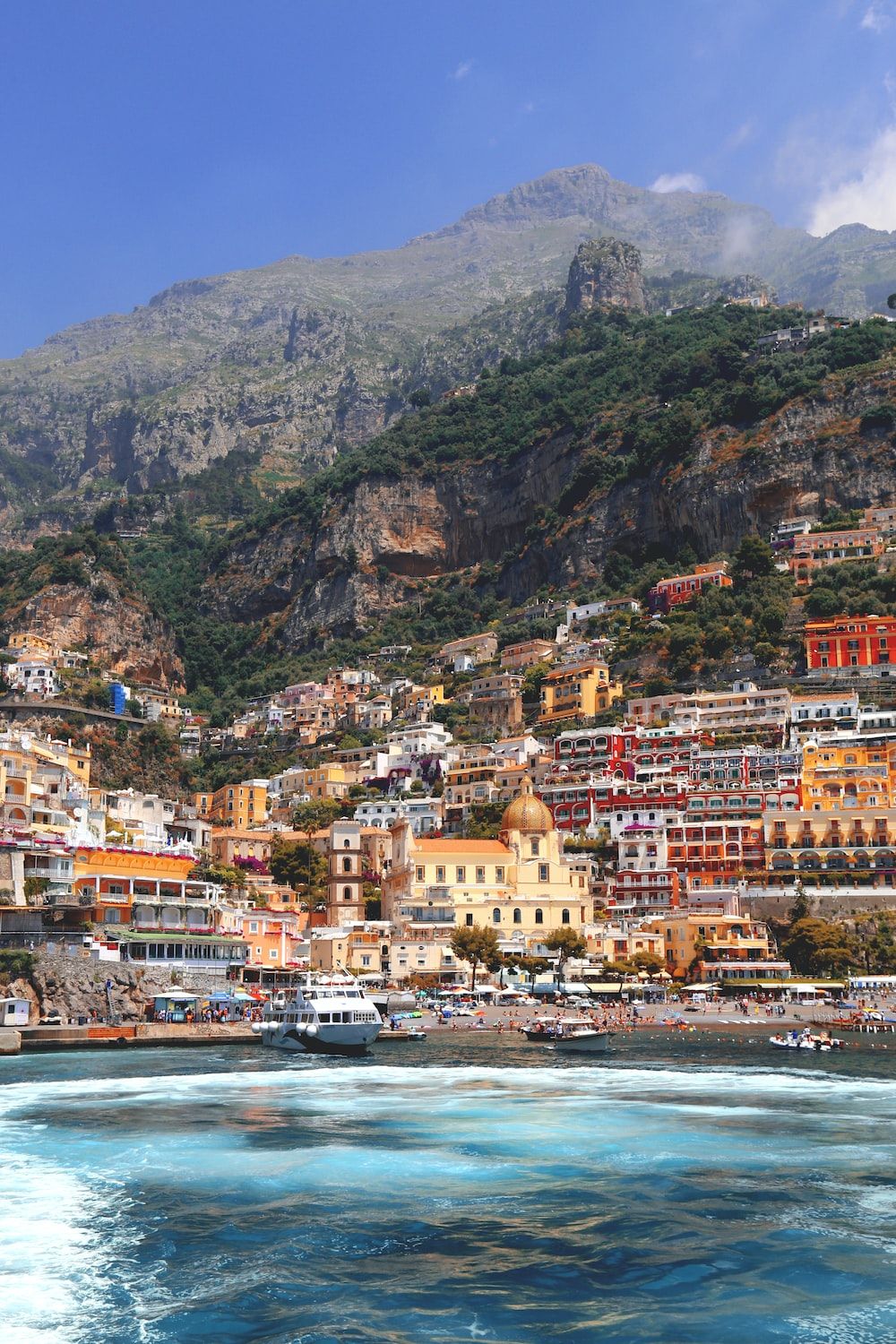 A picturesque town on the coast with colorful buildings and a large mountain in the background. The ocean is in the foreground and blue with white waves. - Italy