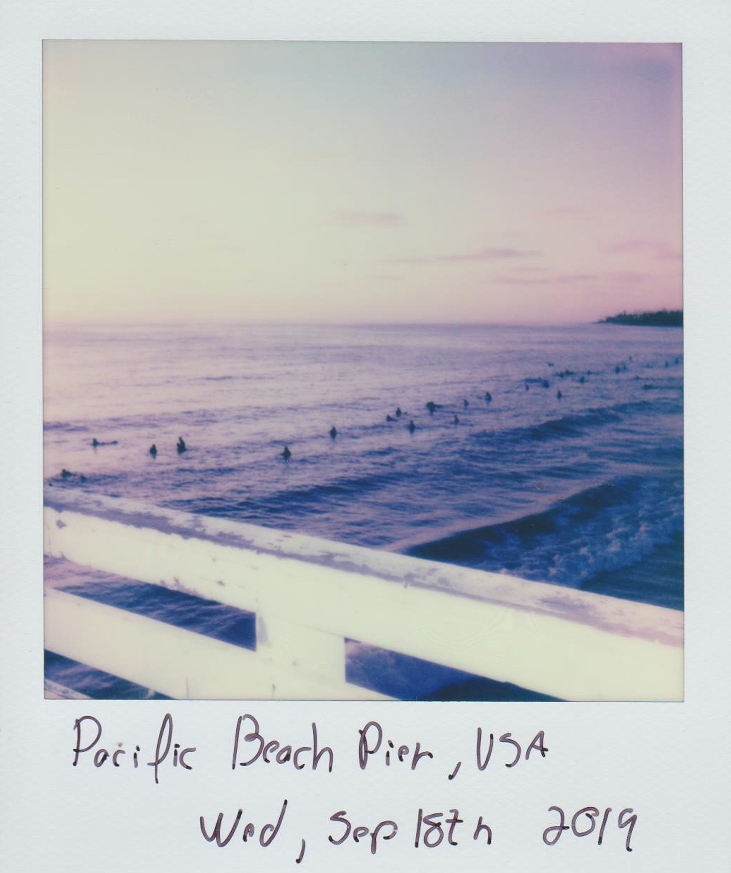 Polaroid of the Pacific Beach Pier in California, USA. The pier is white and has a wooden fence. The ocean is blue and has many surfers in it. The sky is pink. - Polaroid