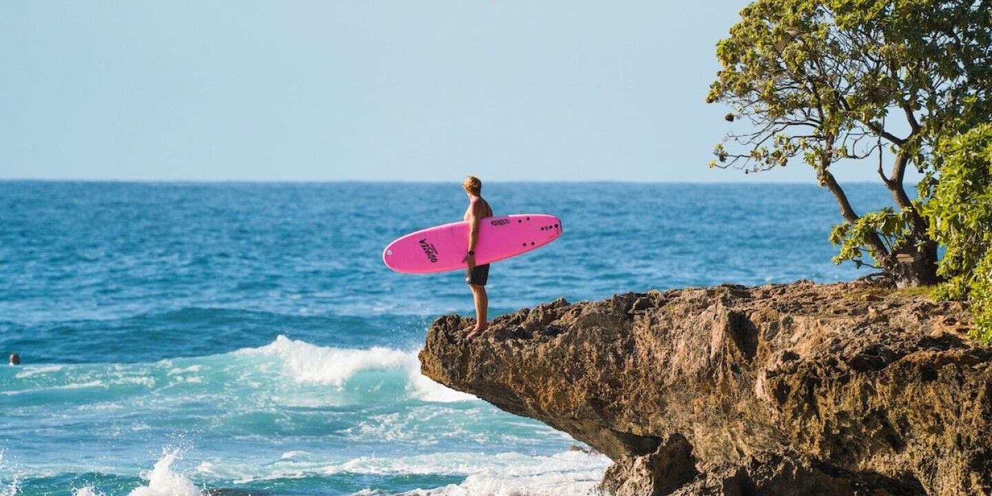 A man stands on a cliff with a pink surfboard, looking out at the ocean. - Surf