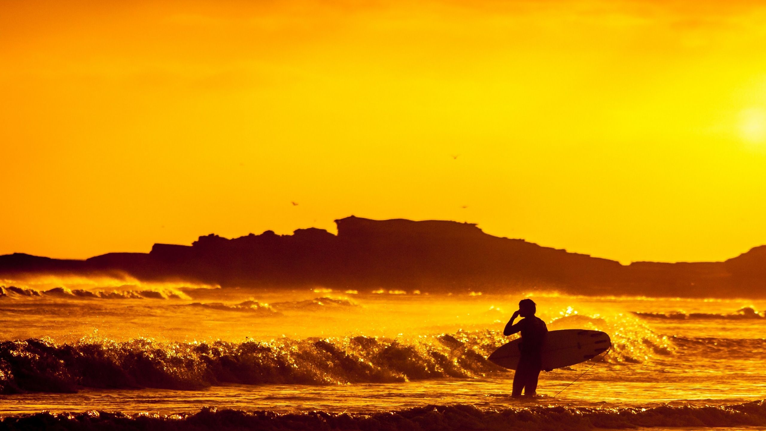 Wallpaper / a surfer stood in the sea at sunset holding a board beneath a vibrant orange dusk sky, orange sunset surfer 4k wallpaper free download