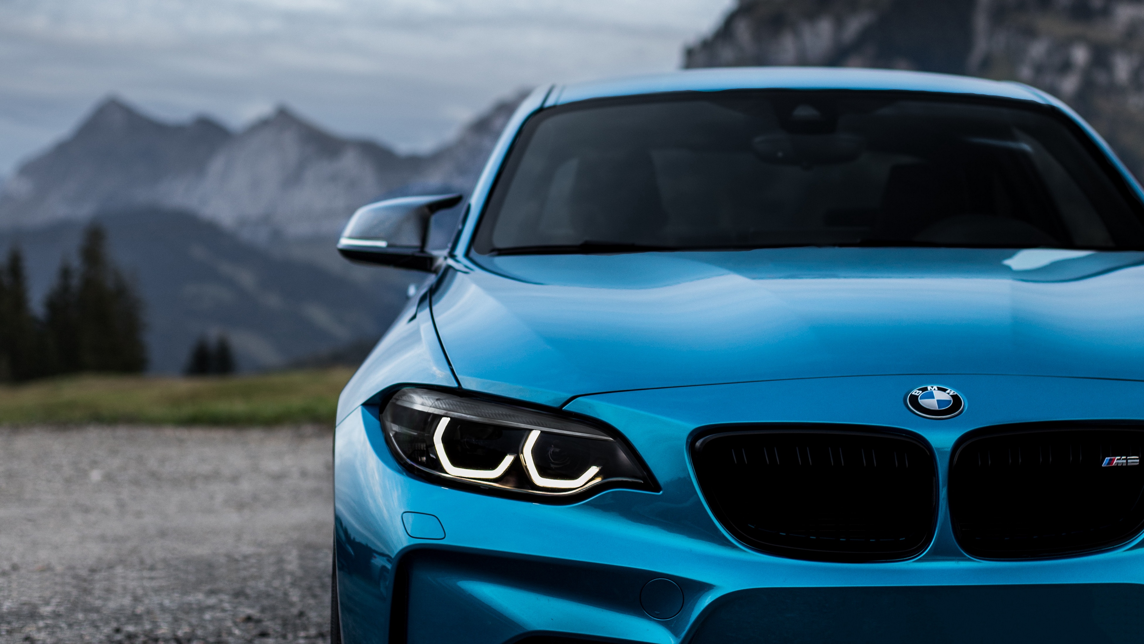 A blue BMW M2 is parked on a gravel road with mountains in the background. - BMW