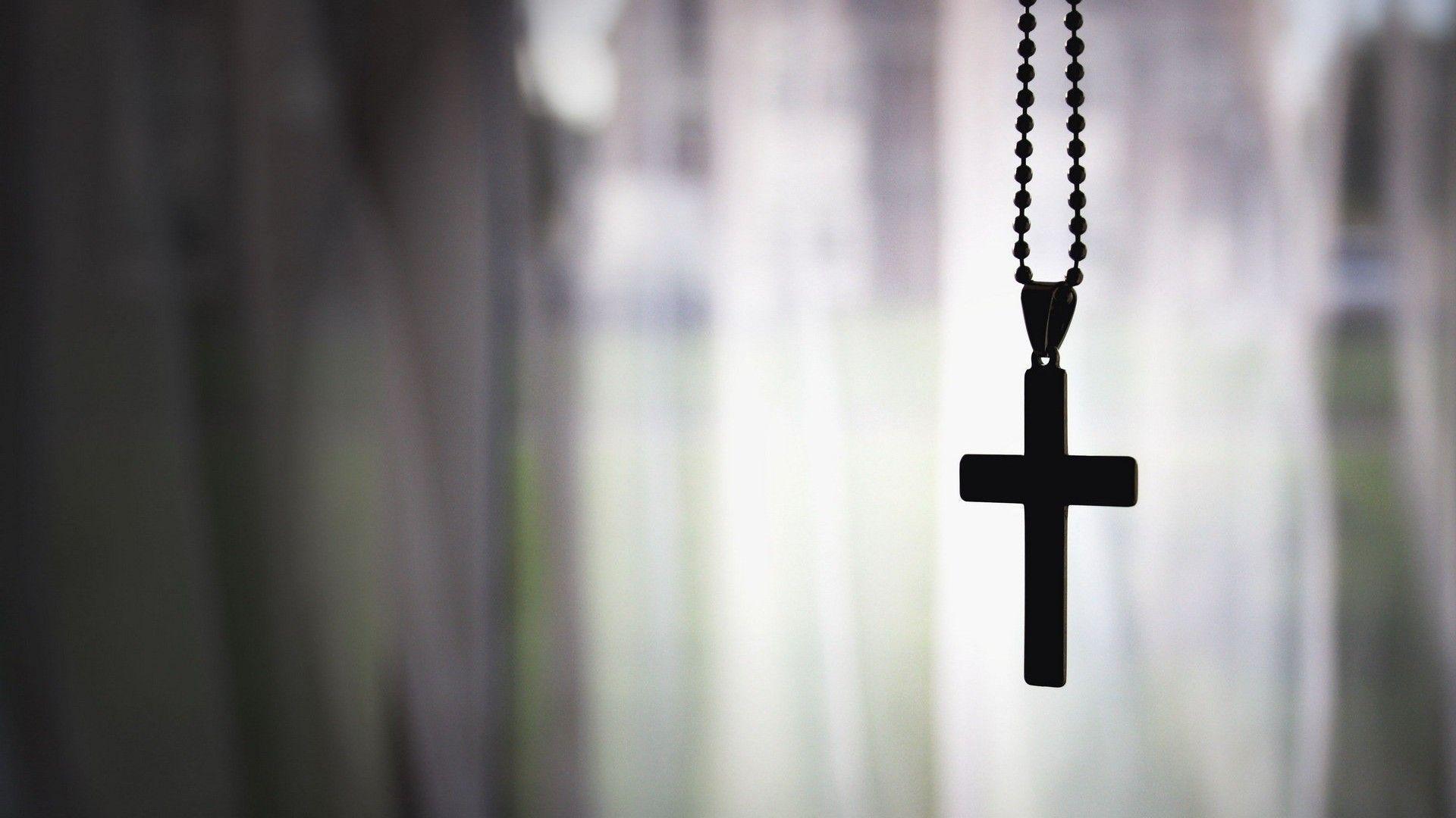 A black cross on a chain hanging in front of a blurred background. - Cross