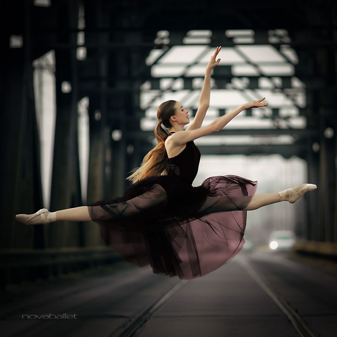 A ballerina in a black dress and white shoes leaps into the air with her arms raised and her legs split. - Ballet