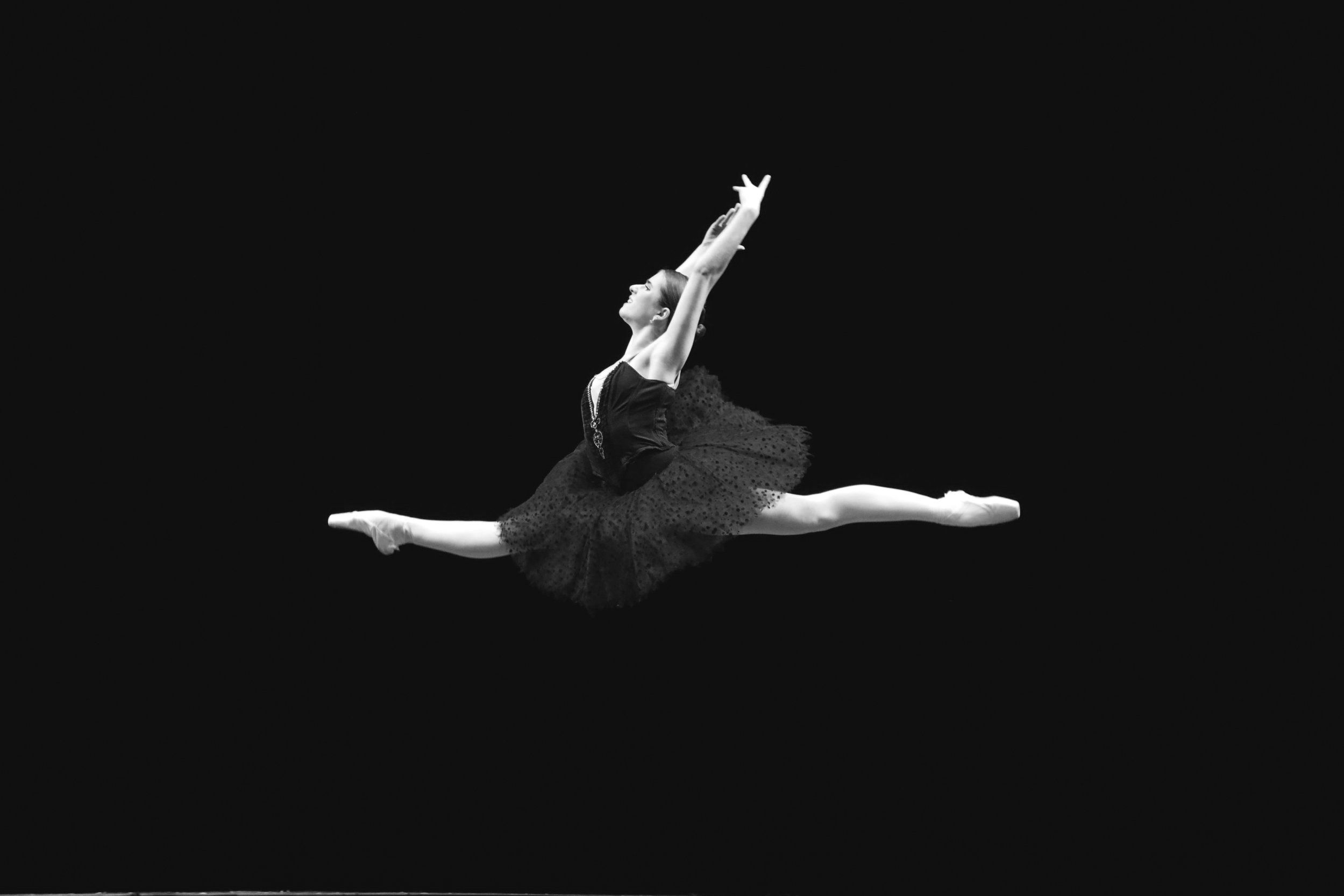 A ballerina in a black tutu leaps into the air with her arms raised. - Ballet