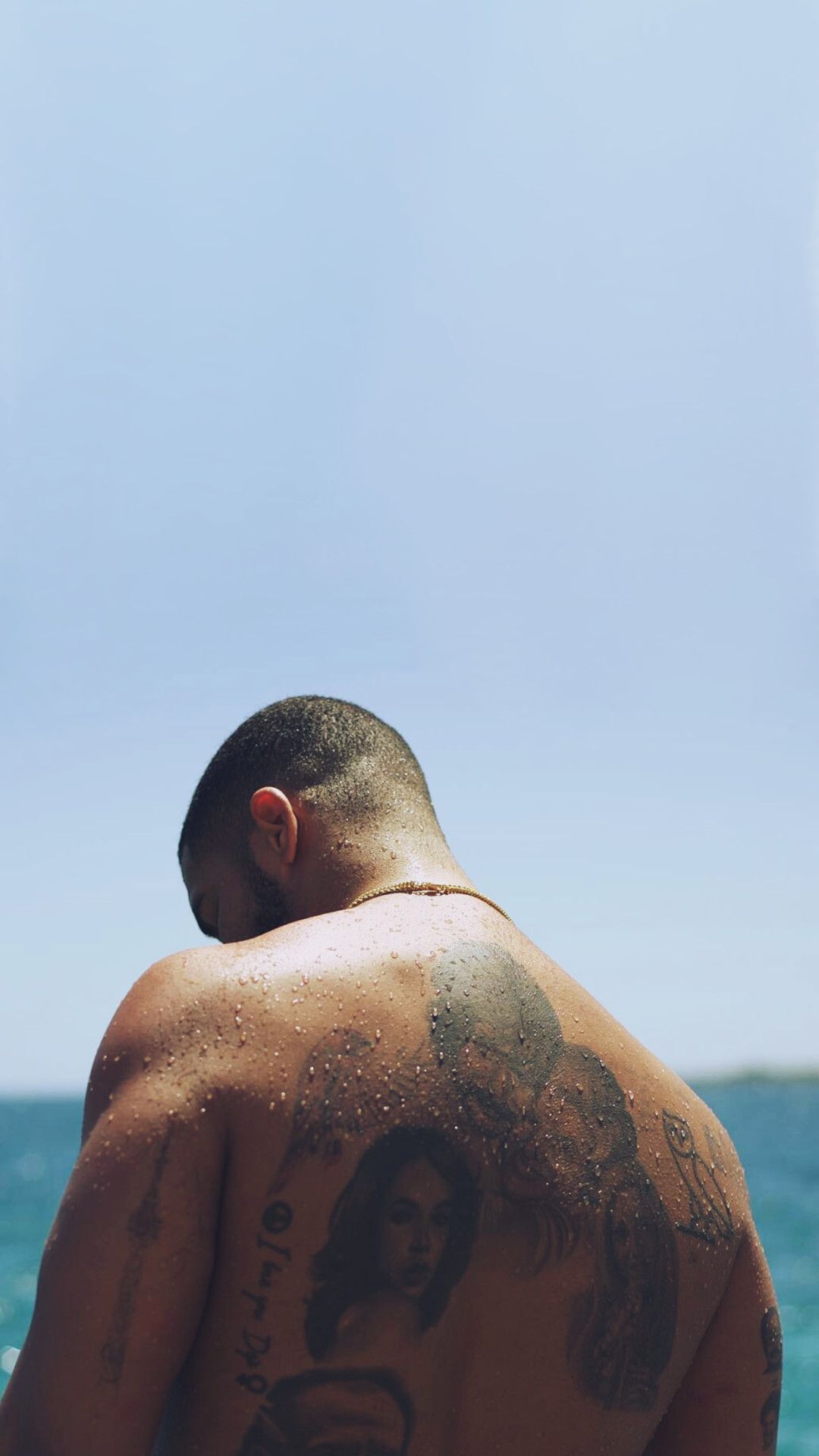 A man with tattoos on his back standing on the beach - Drake
