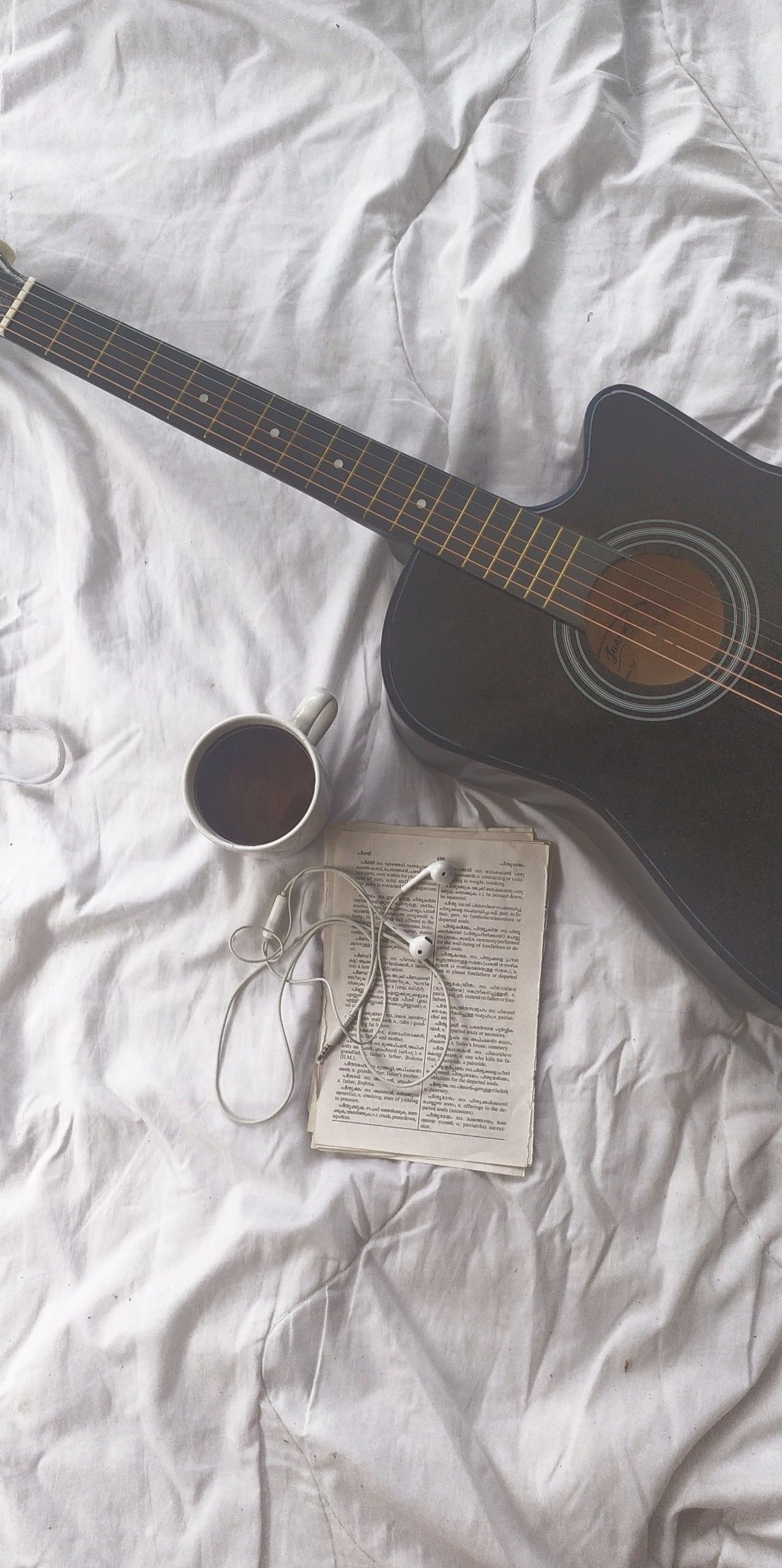 A guitar, a cup of coffee, and some earphones on a bed. - Guitar