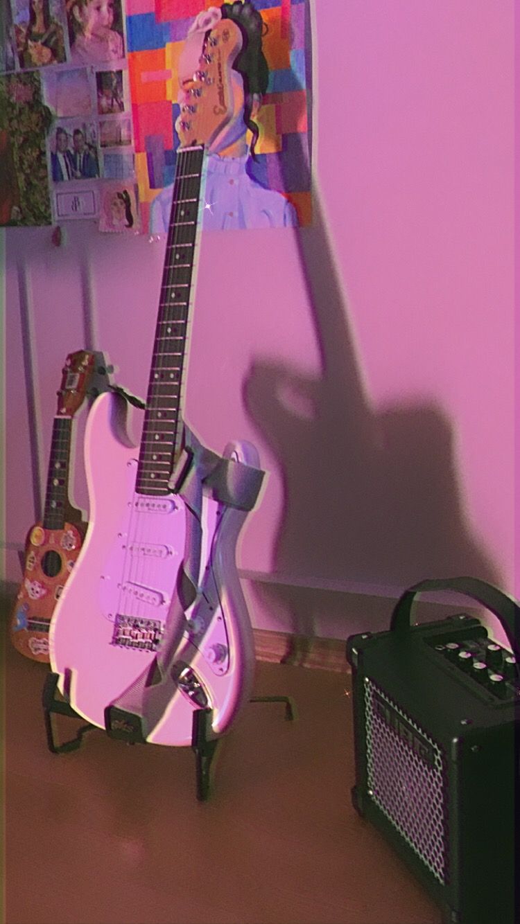 A pink guitar is propped up against a wall next to a black speaker. - Guitar