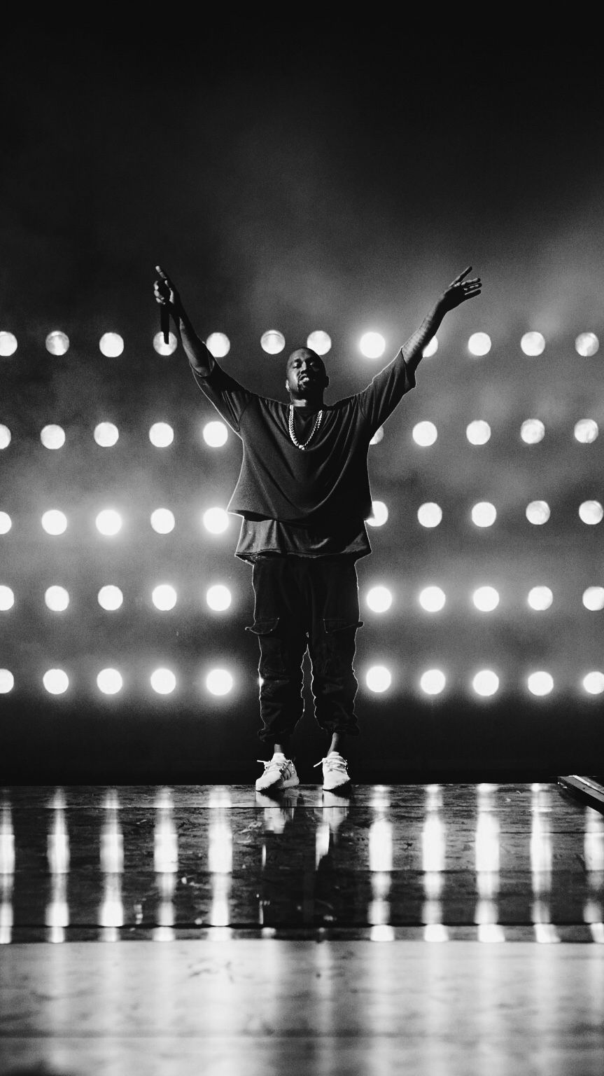 A black and white photo of Kanye West on stage with his arms raised in the air. - Kanye West