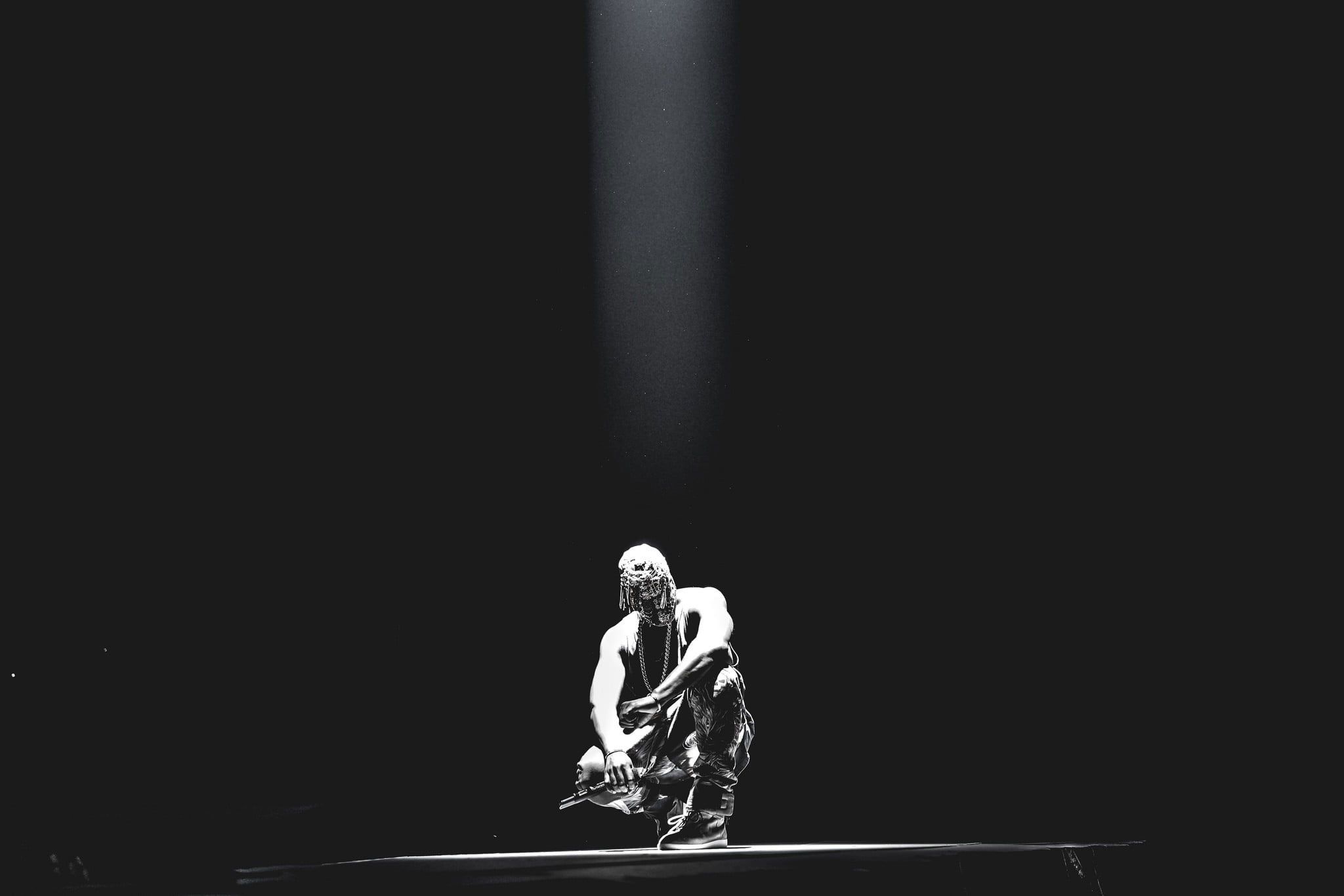 A man in a white tank top and camouflage pants crouches on the floor in the spotlight. - Kanye West