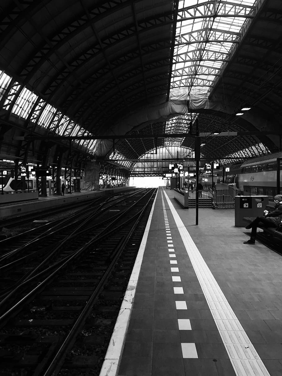 A black and white photo of a train station with a person sitting on a bench. - Black and white