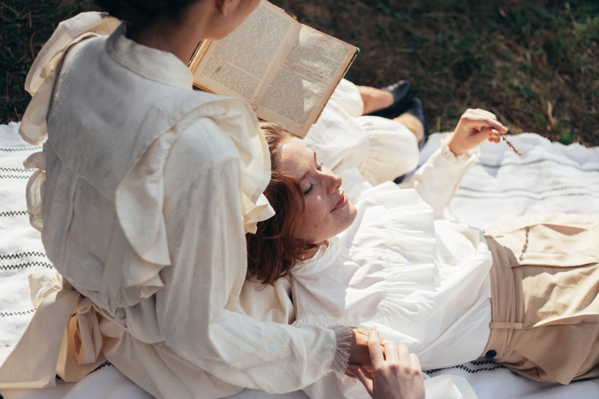 Two women in white lay on a blanket with one reading a book - Light academia
