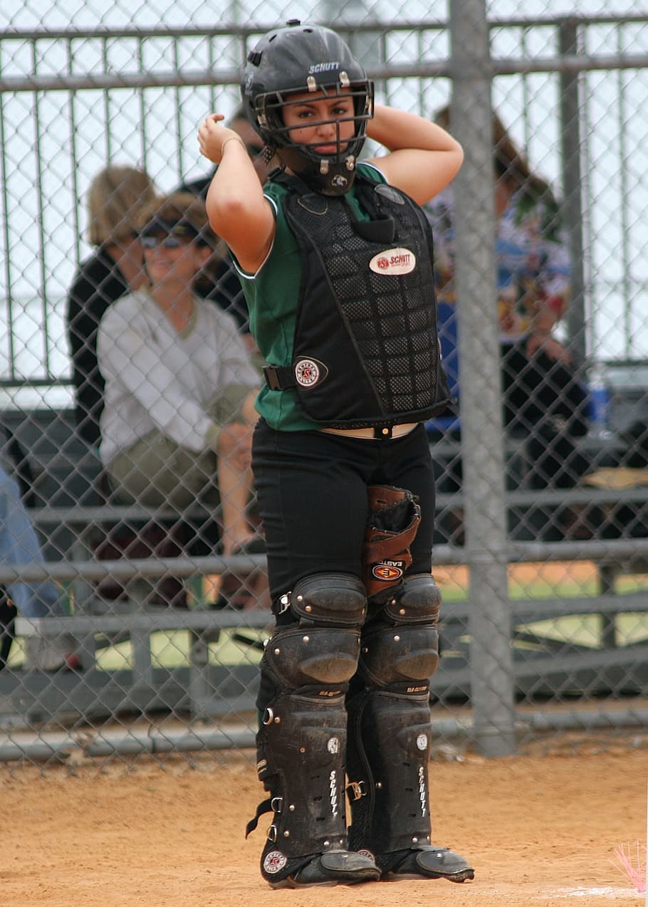 A girl in a baseball uniform and catcher's gear is standing on a field. - Softball