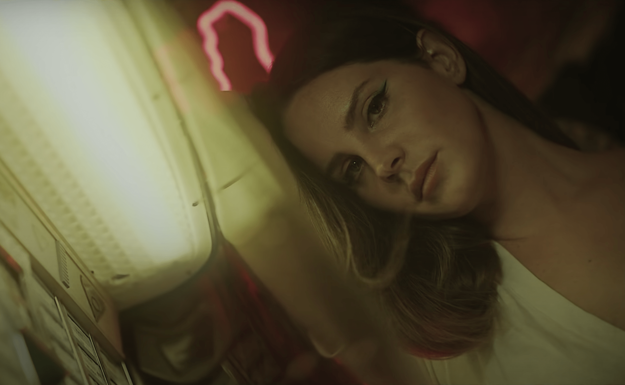 A woman laying on a bed with a neon sign in the background - Lana Del Rey