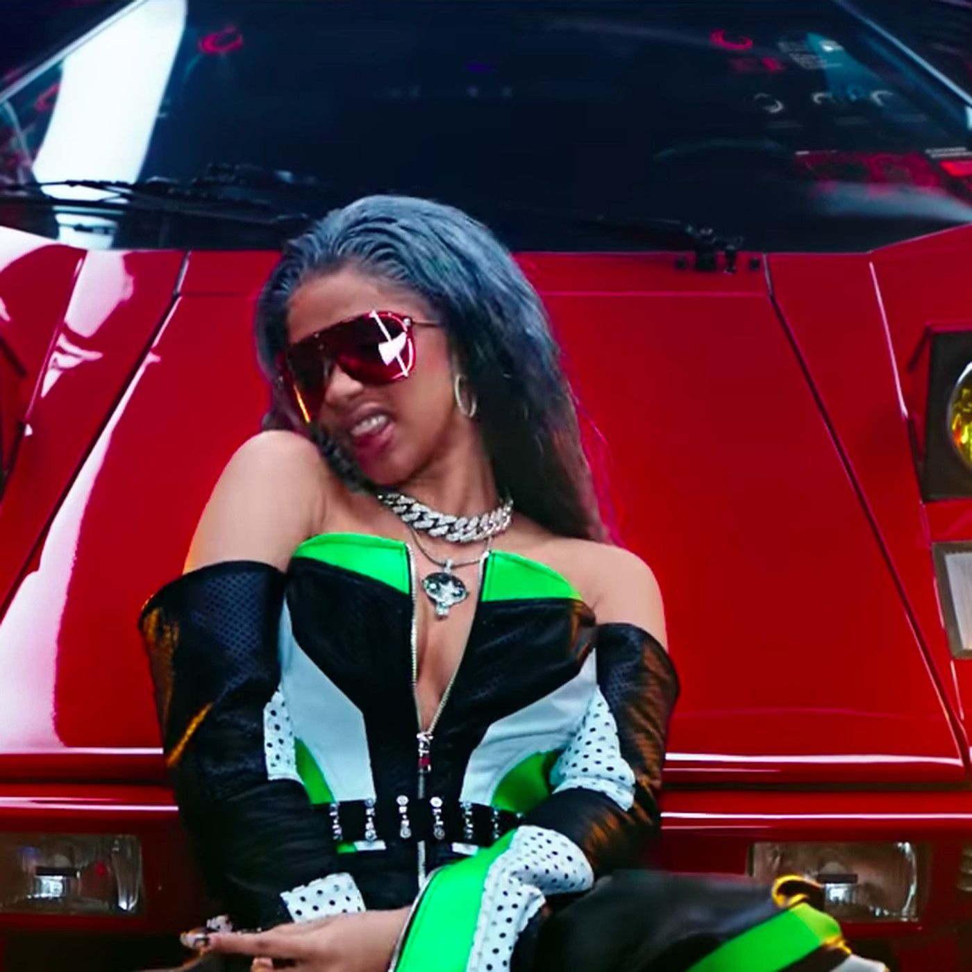 Cardi B sitting on the hood of a red car in a black and green top and sunglasses - Cardi B