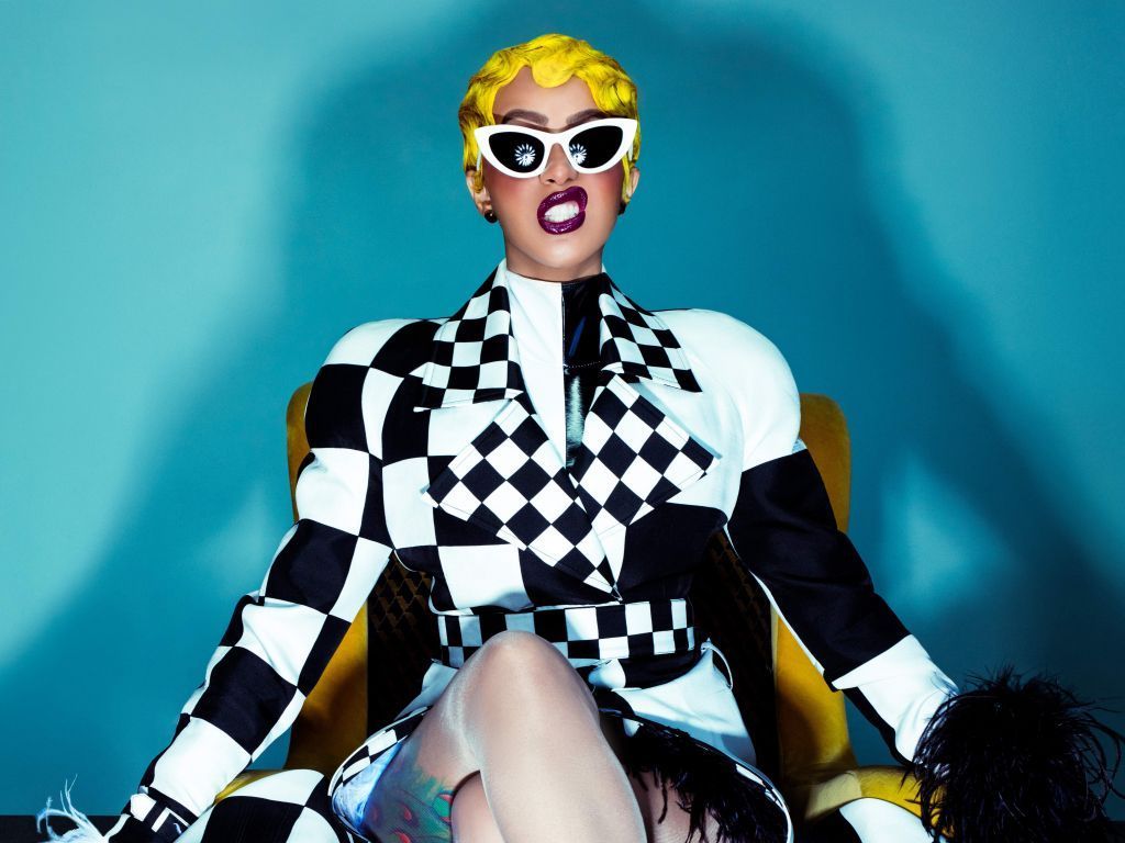 A woman with yellow hair and white sunglasses sits on a chair. - Cardi B