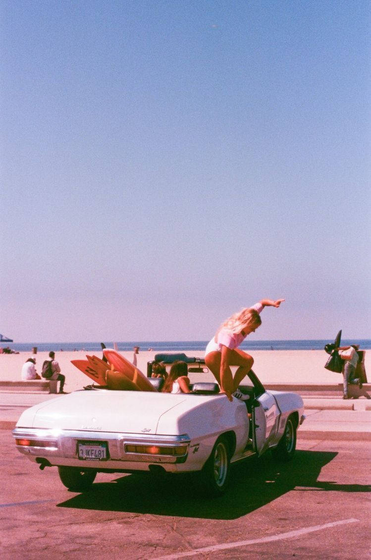 A woman in a white bathing suit does a cartwheel on the hood of a white convertible. - 90s