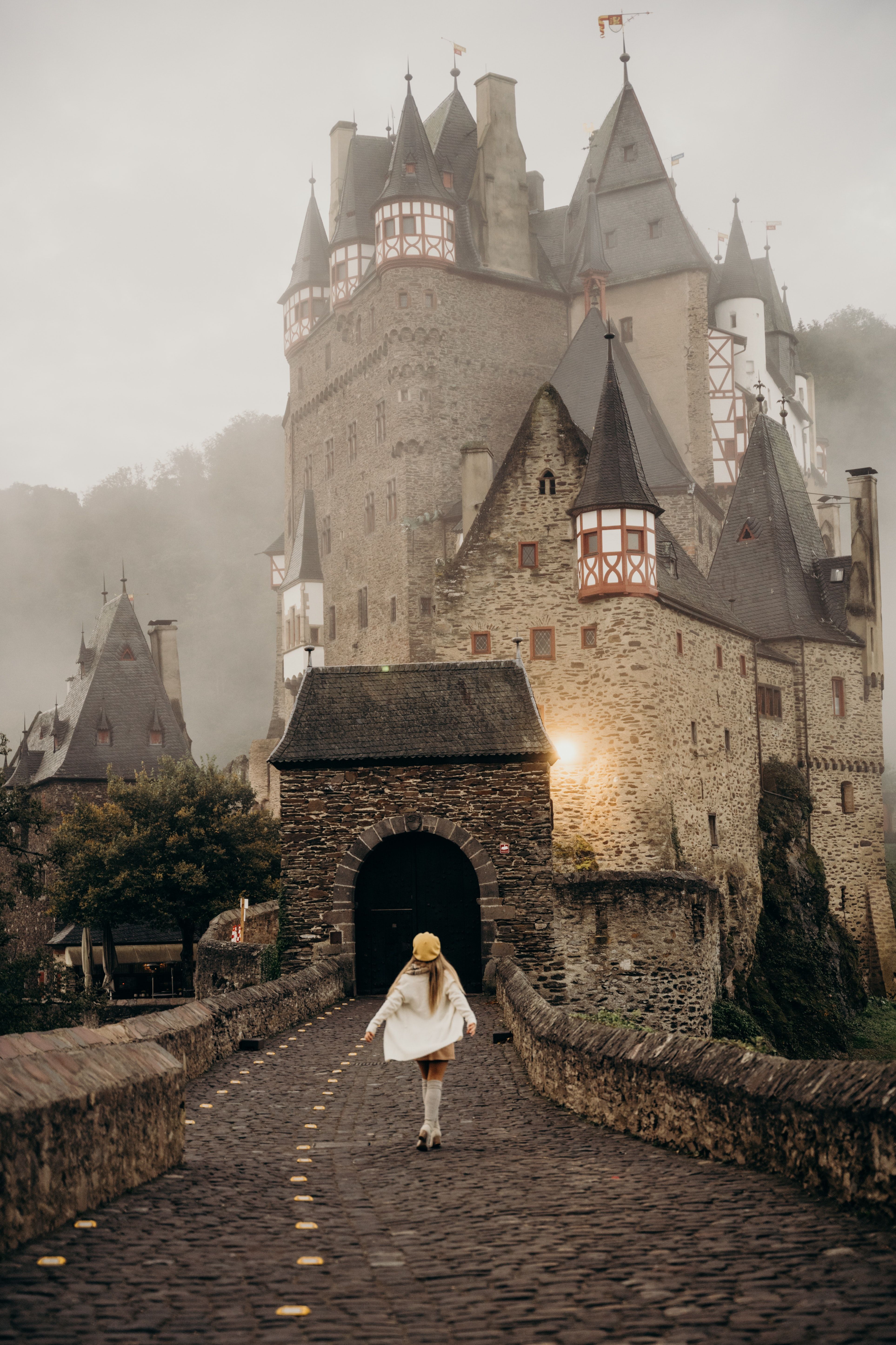 A woman in a white coat and yellow hat walks towards a castle in the fog. - Castle
