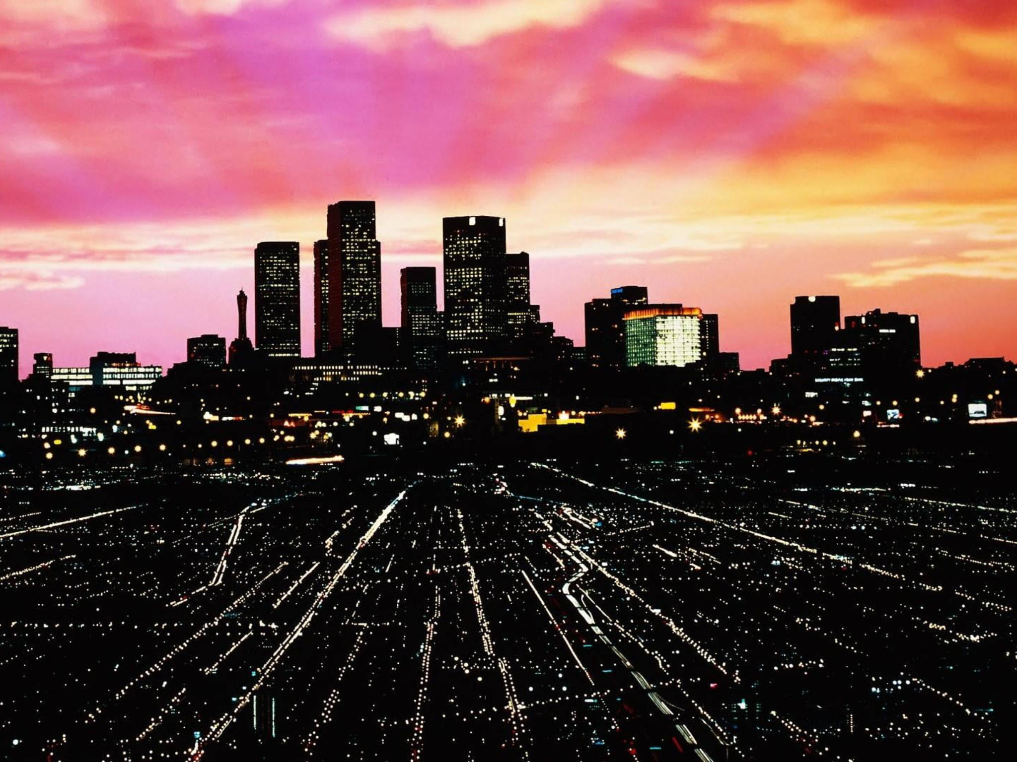 This image is a photo of a city skyline during sunset. The city skyline has multiple buildings of varying heights, and the photo is taken from a distance that includes a large highway filled with traffic. The sky is a mix of purple, pink, and orange colors due to the sunset. - Los Angeles