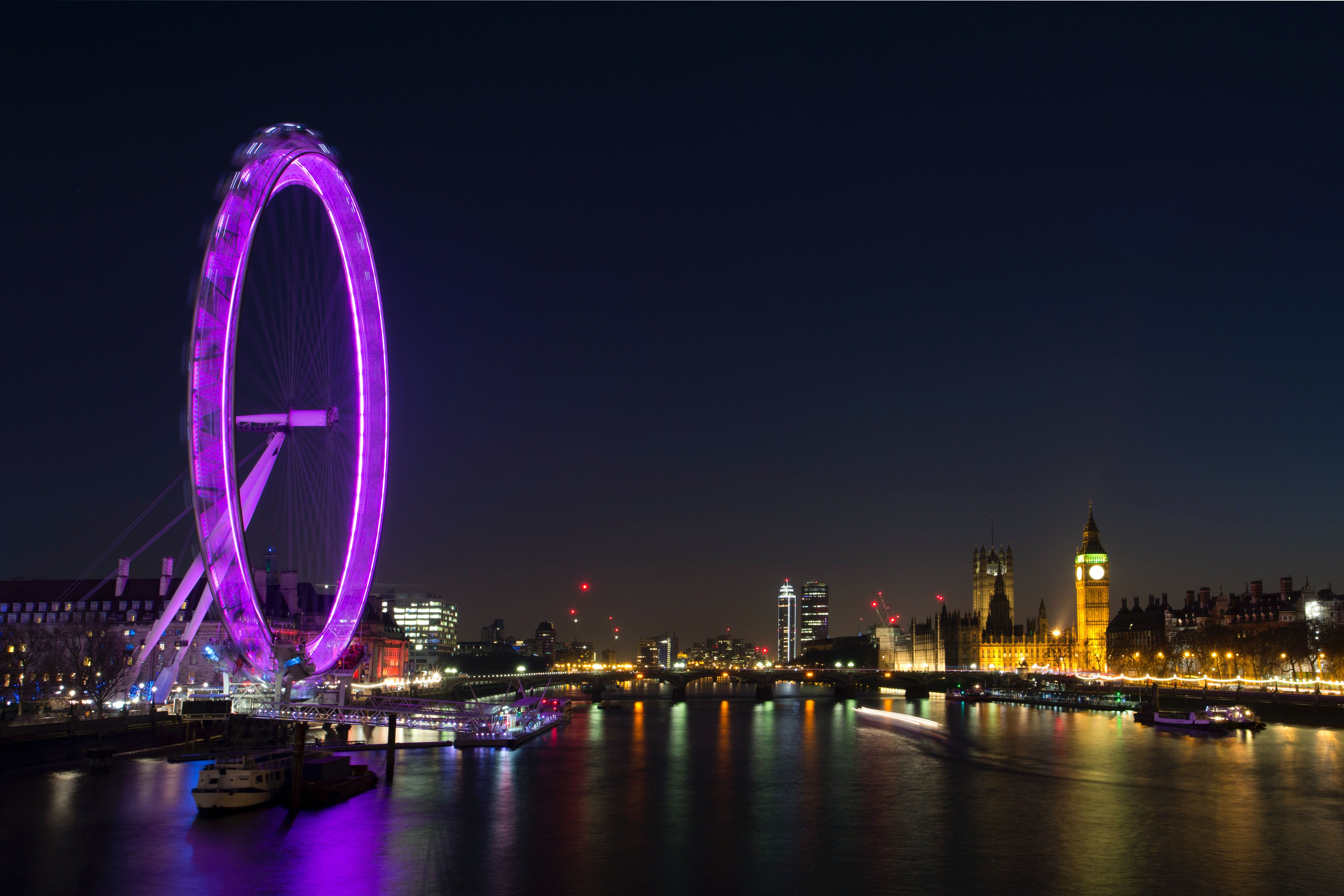 A purple lit up wheel over looking a river with boats in it. - London