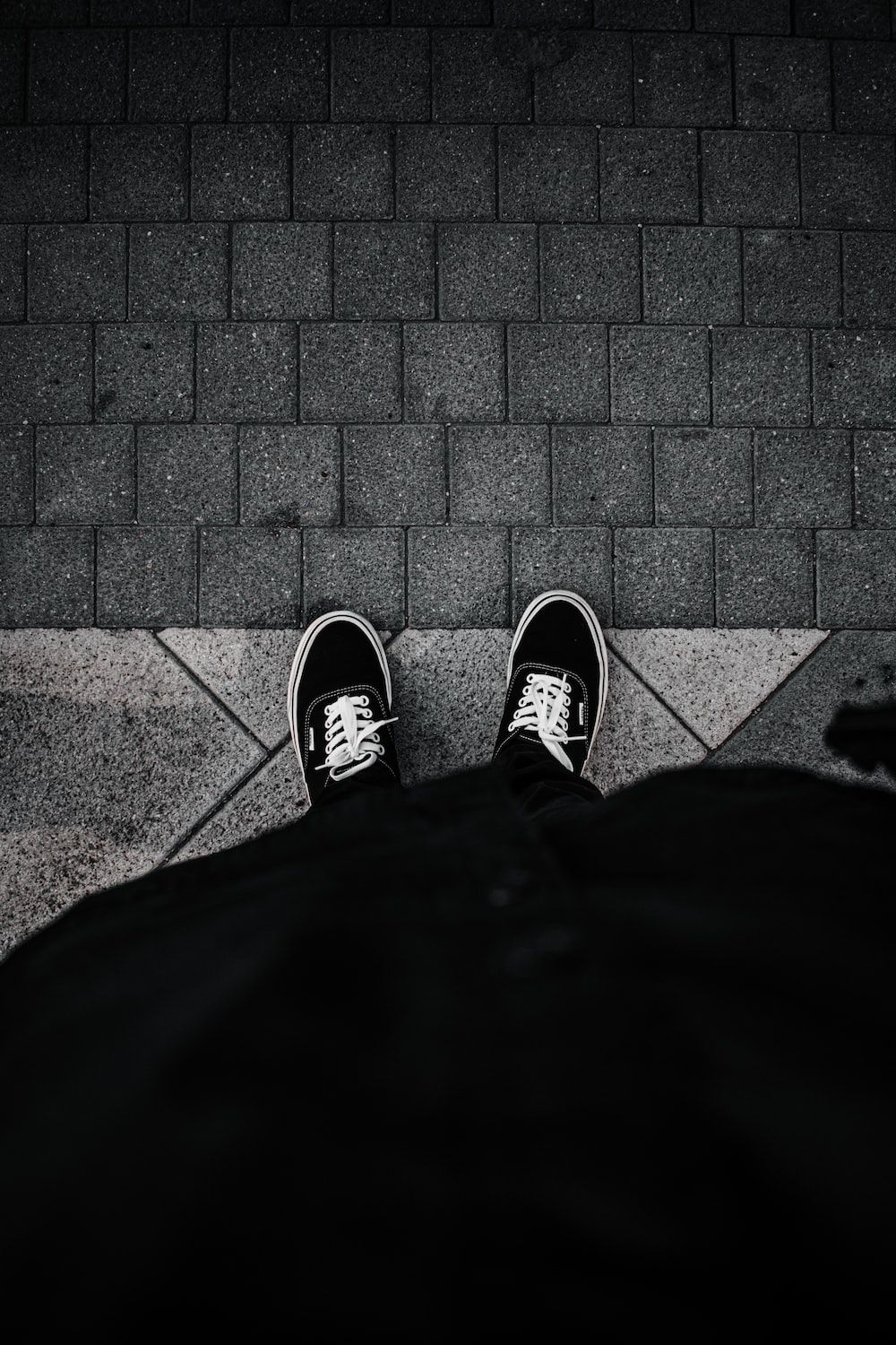 A person wearing black and white Vans sneakers standing on a sidewalk - Vans