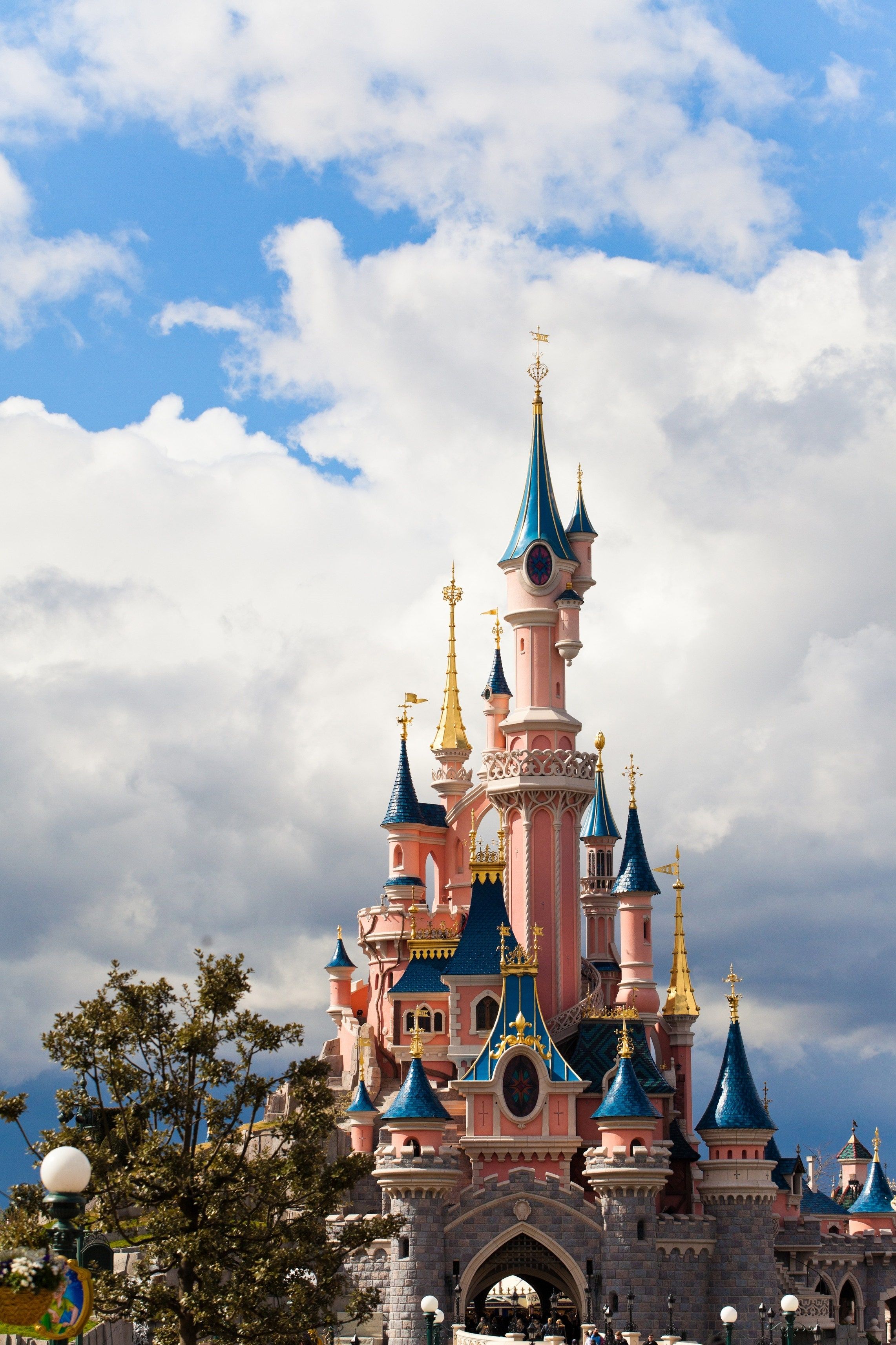 A pink castle with blue and gold spires stands in front of a blue sky with white clouds. - Disneyland