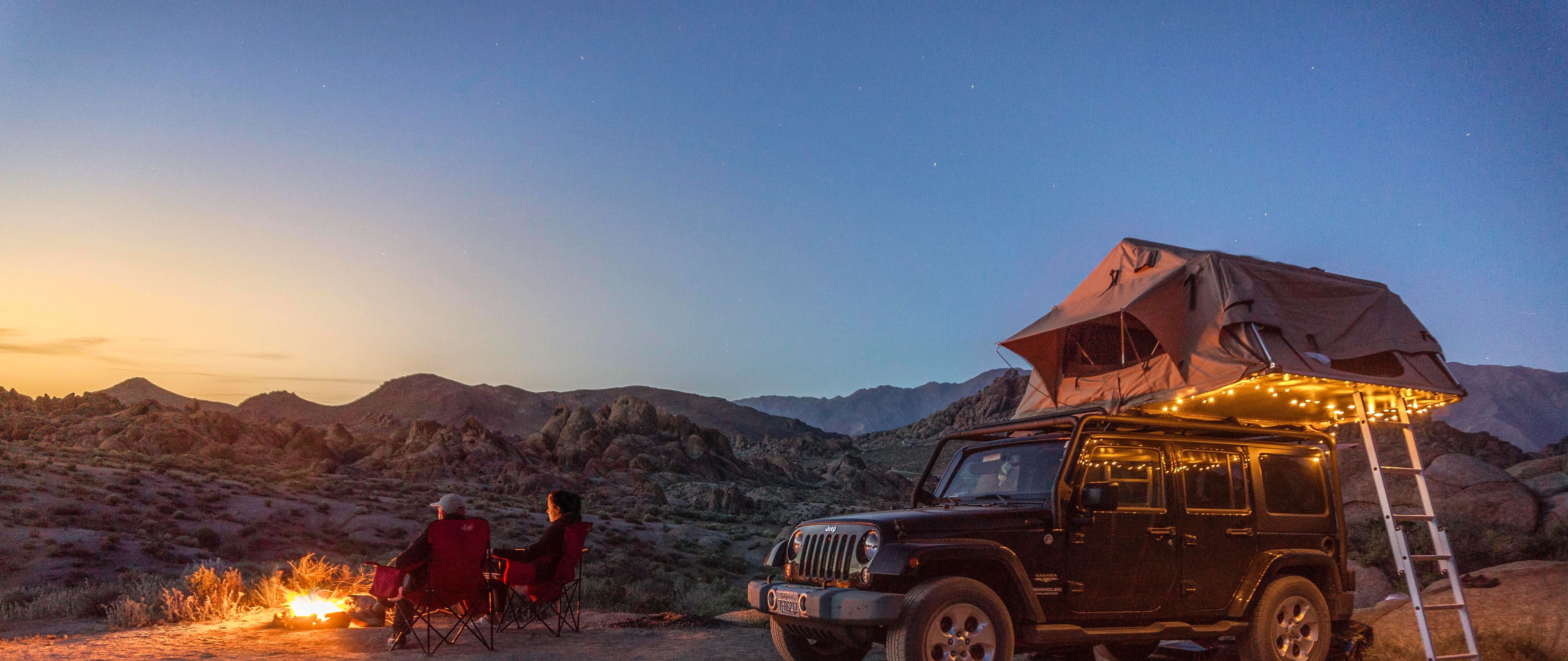 A jeep with a tent on top parked in front of a campfire in the mountains. - Camping