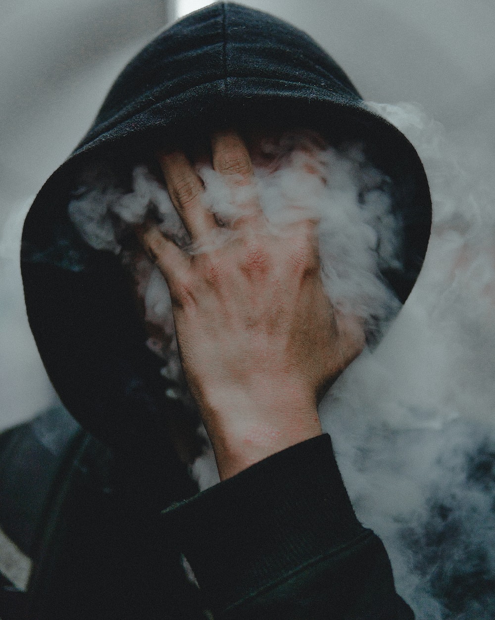 A person in a black hoodie covers their face with their hand as smoke billows around them. - Pop Smoke