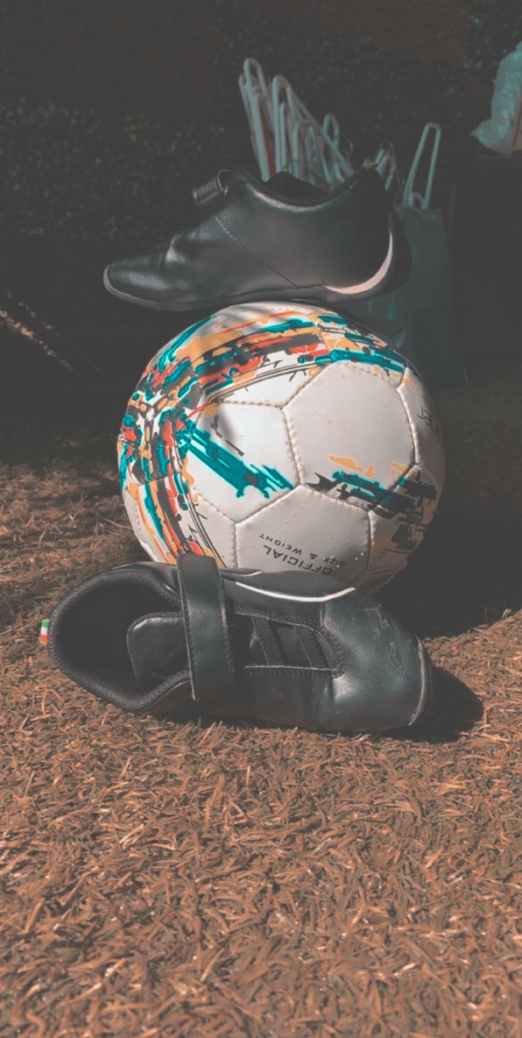 A soccer ball sits on the ground next to a pair of black soccer cleats. - Soccer