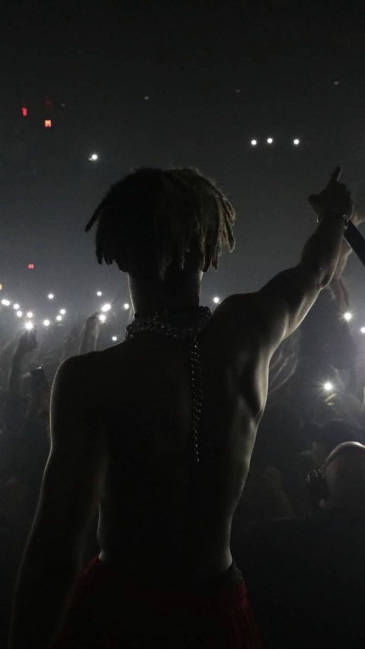 A shirtless man with dreads stands in front of a crowd with his arm raised. - XXXTentacion