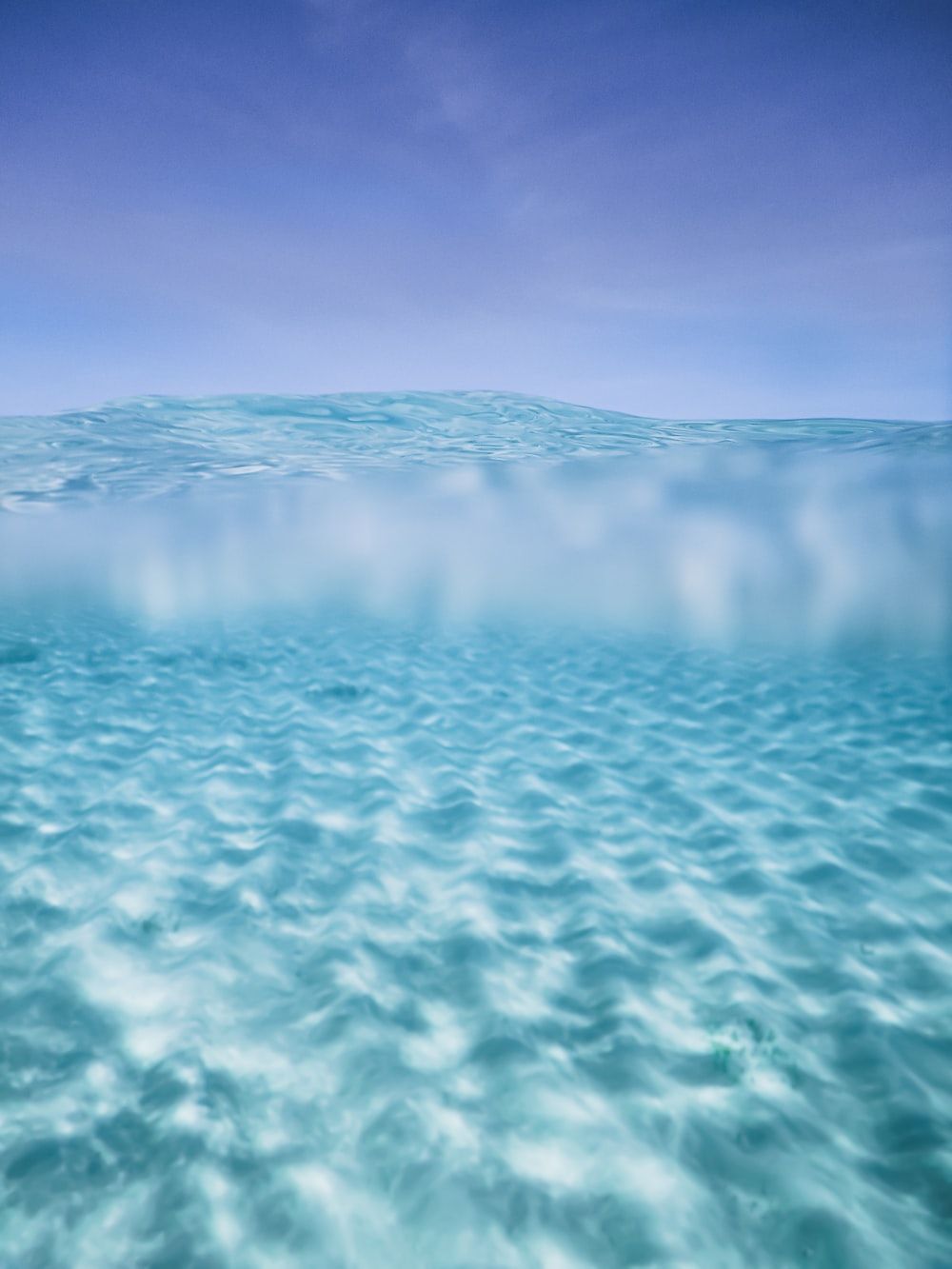This image is a painting of an ocean scene, divided in half. The top half is clear blue sky, while the bottom half is a gradient of blue to white, representing the ocean floor. The water is depicted with ripples on the surface. - Underwater