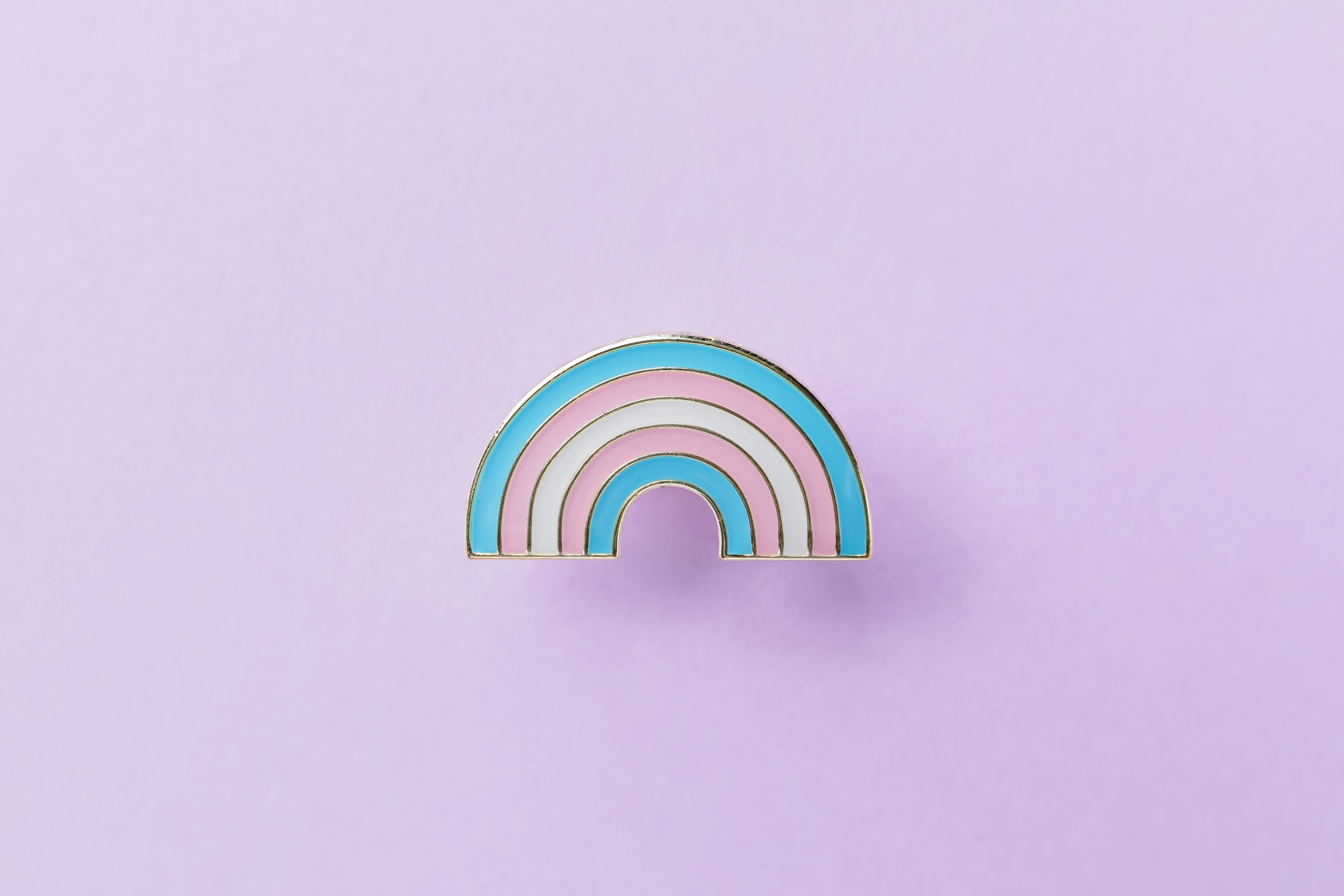 A transgender pride pin in the shape of a rainbow, on a purple background. - Non binary