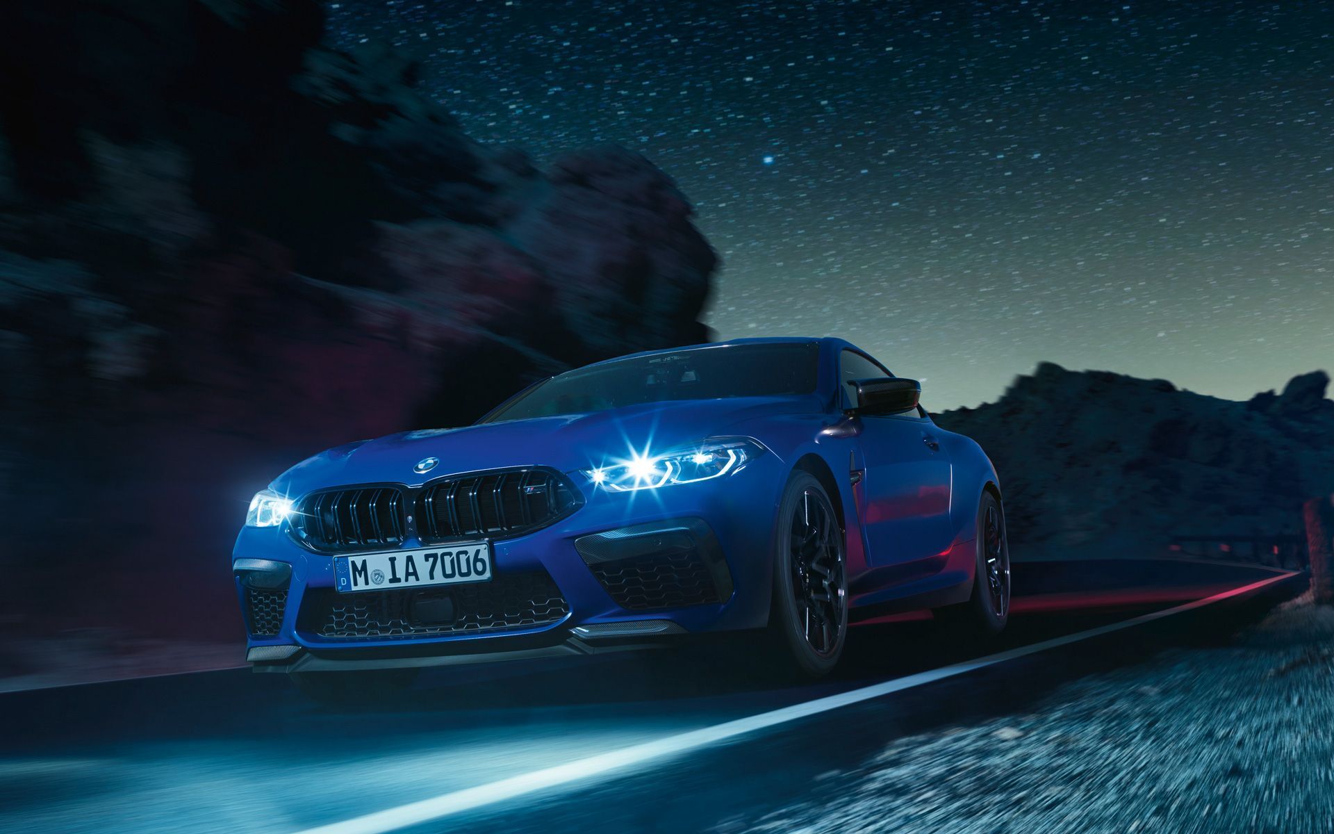 The BMW M8 Competition Coupé in Yas Marina Blue metallic driving on a road at night with lights on. - BMW
