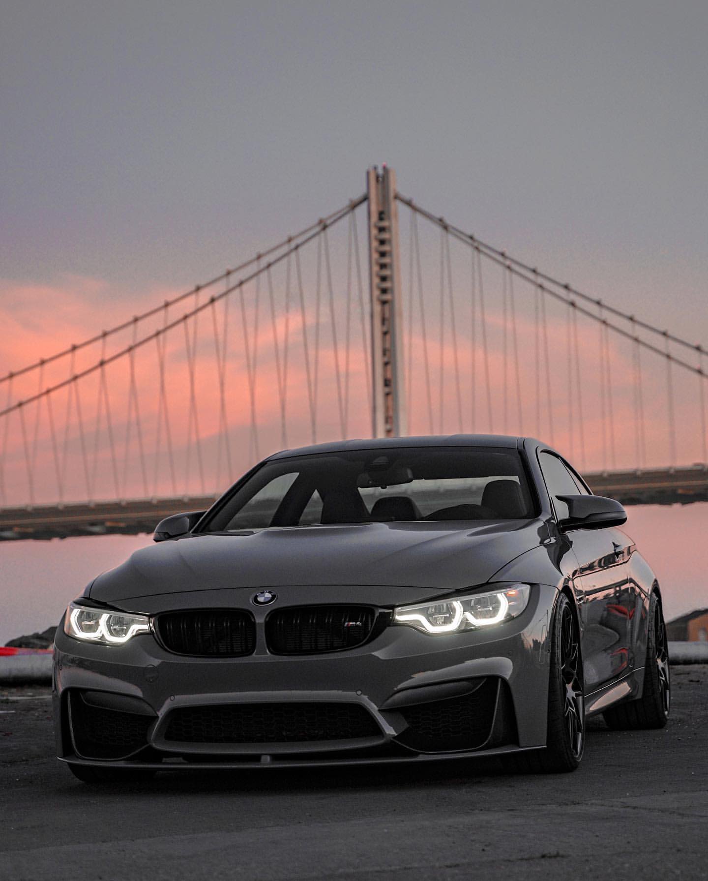 A grey BMW M4 with a front lip from Eibsee is parked in front of a bridge with the sunset in the background. - BMW