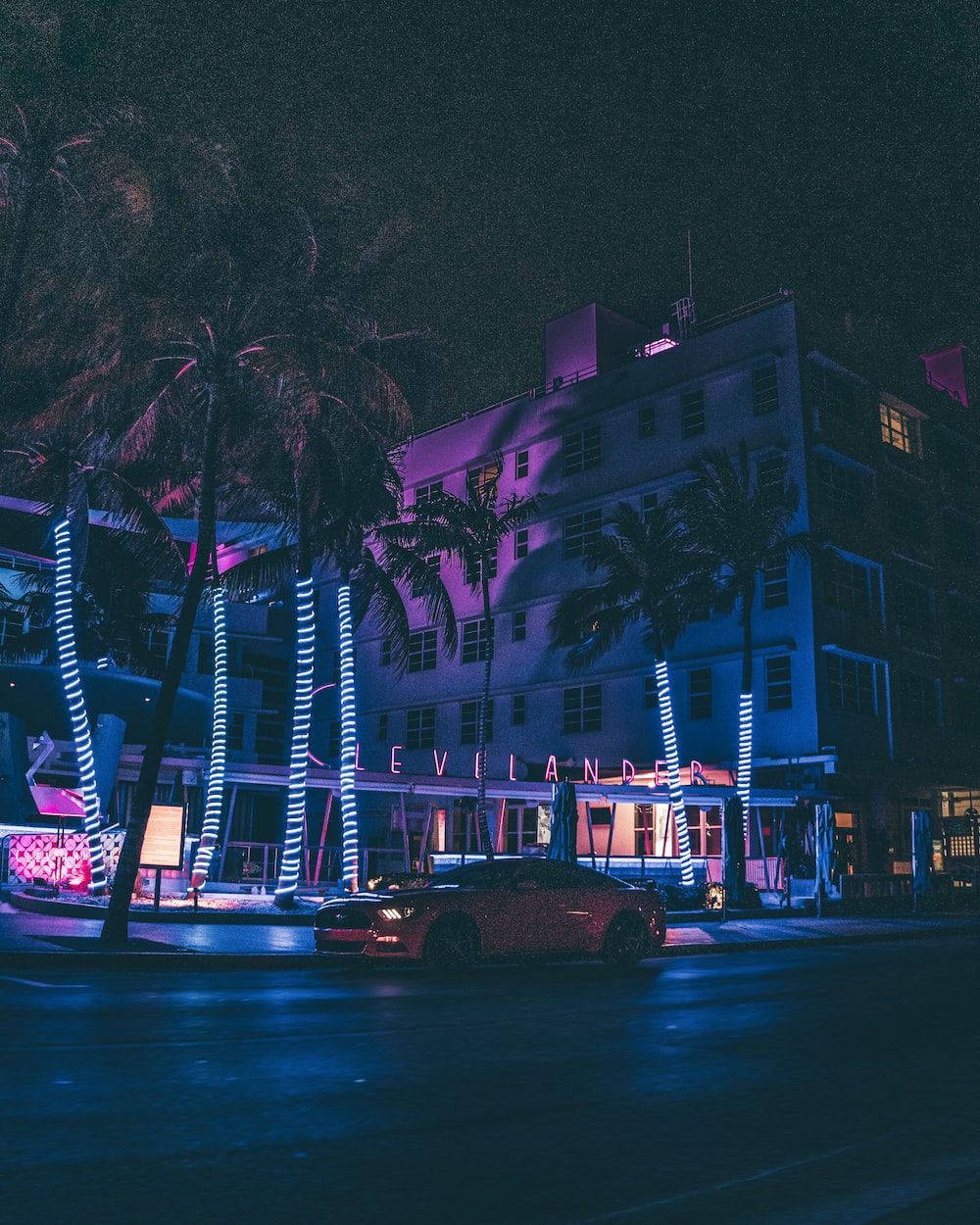 A city street at night with neon lights on the buildings and palm trees. - Miami