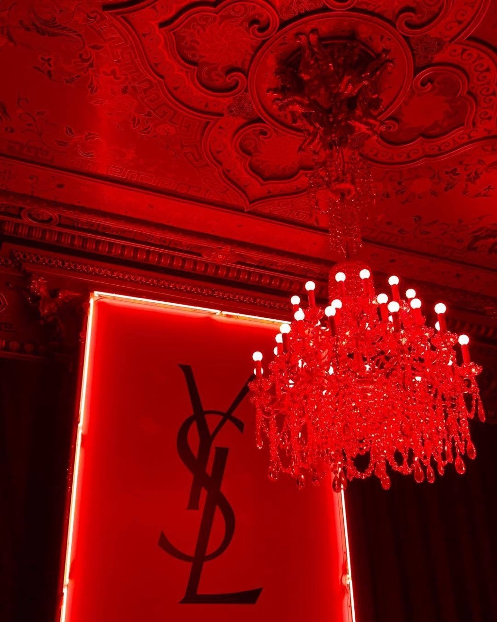 A red chandelier hangs from a red ceiling with a red YSL sign below it. - Light red