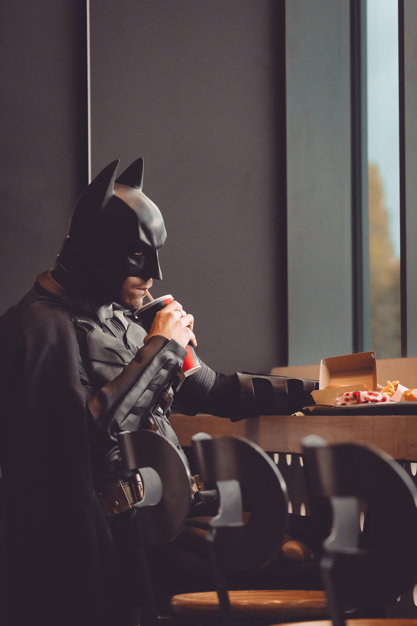 A man in a Batman costume sits at a table with a drink and a pizza. - Batman