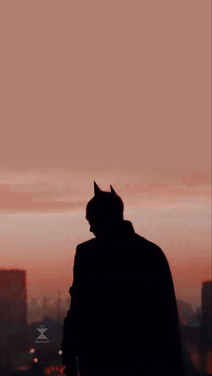 A silhouette of a man in a Batman costume looking out over a city at sunset. - Batman