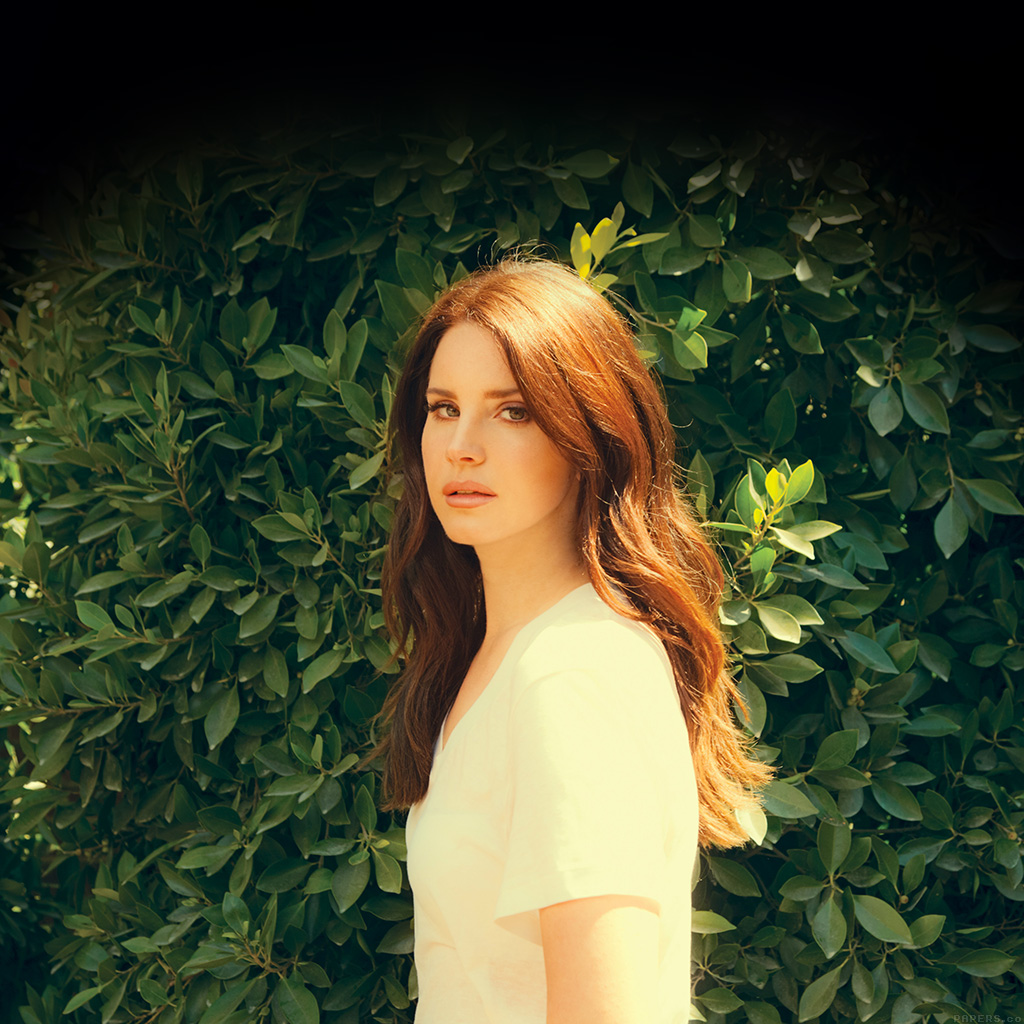 A woman with long red hair standing in front of a wall of green leaves - Lana Del Rey