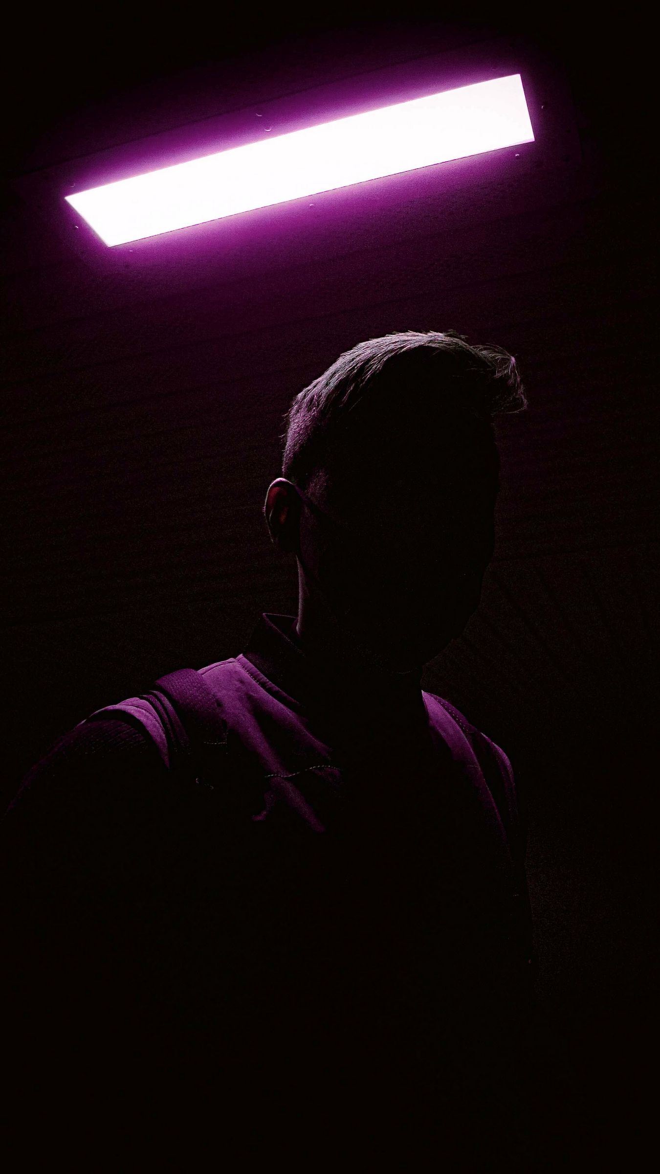 A man standing under a purple neon light in a dark room with his back turned. - Shadow