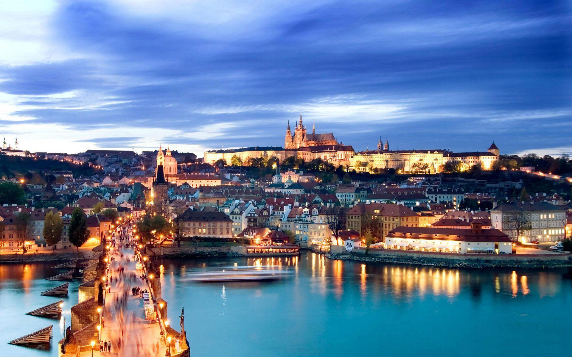 This is a breathtaking view of the city of Prague, taken at dusk. The Vltava River winds through the city, with the Charles Bridge stretching across it. The city is lit up, highlighting the beautiful architecture. In the distance, the Prague Castle sits atop the city, overlooking all below. The sky is a mix of blue and purple, with wispy clouds adding to the overall beauty of the scene. - 1920x1200