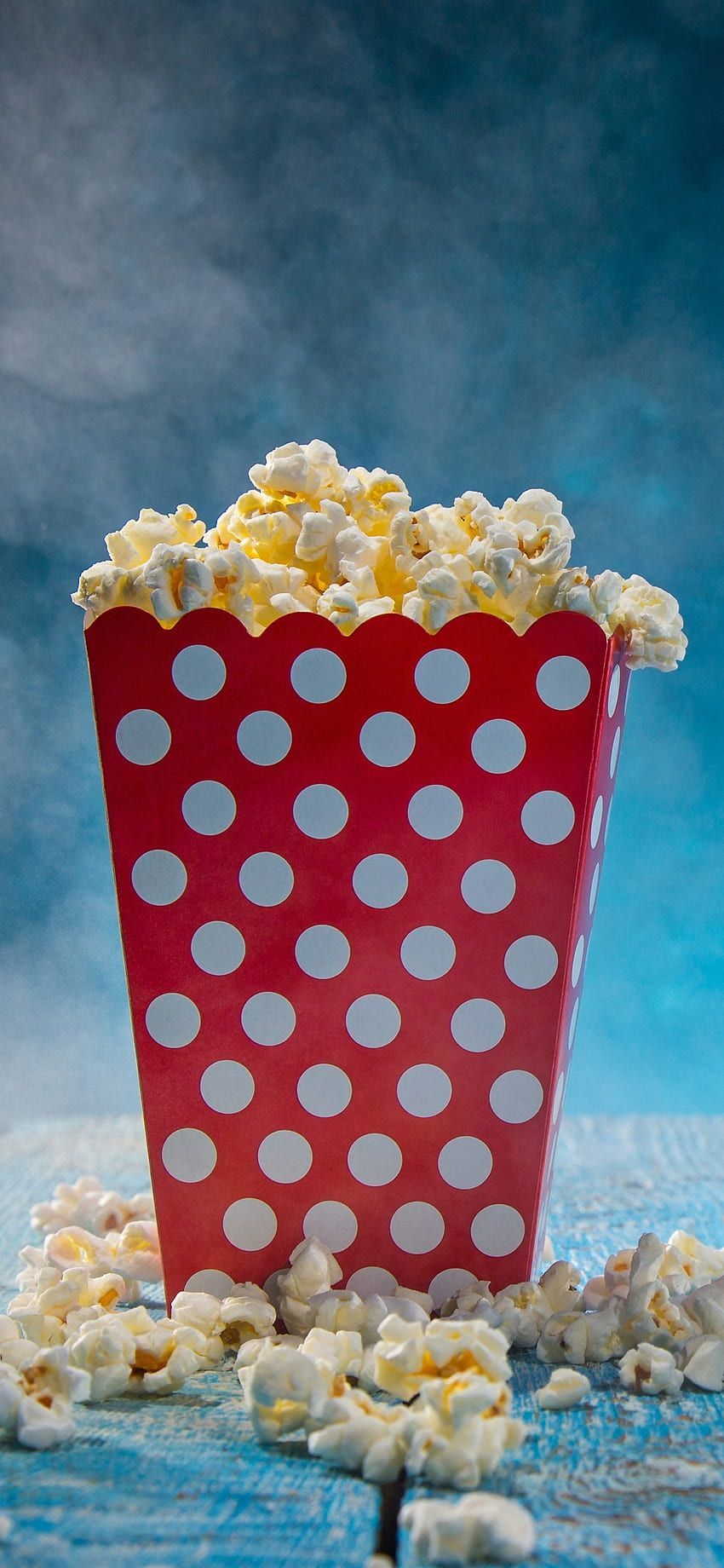 A red and white polka dot popcorn bucket overflowing with popcorn on a blue background - Popcorn