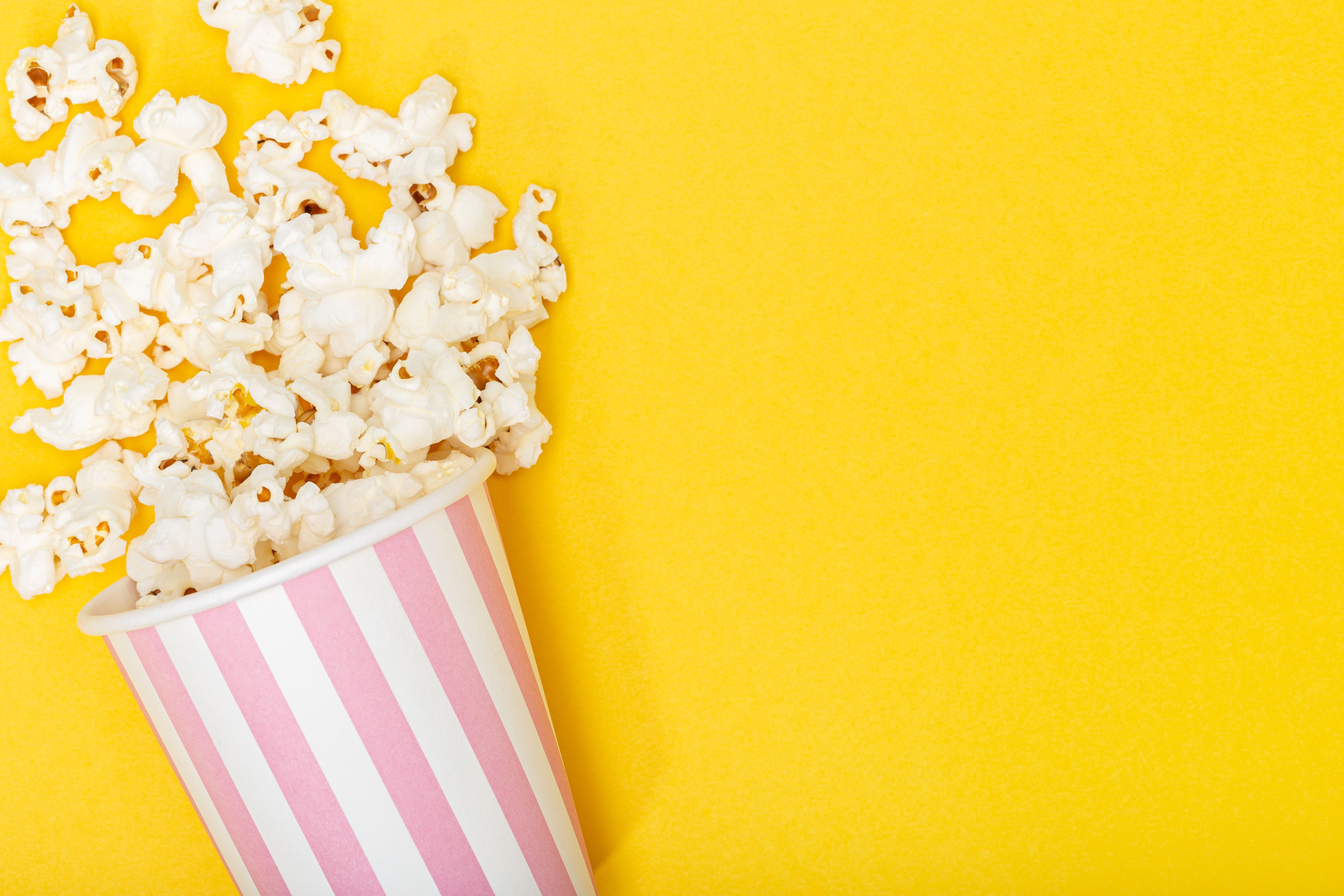 A pink and white striped popcorn bucket with popcorn spilling out of it on a yellow background. - Popcorn