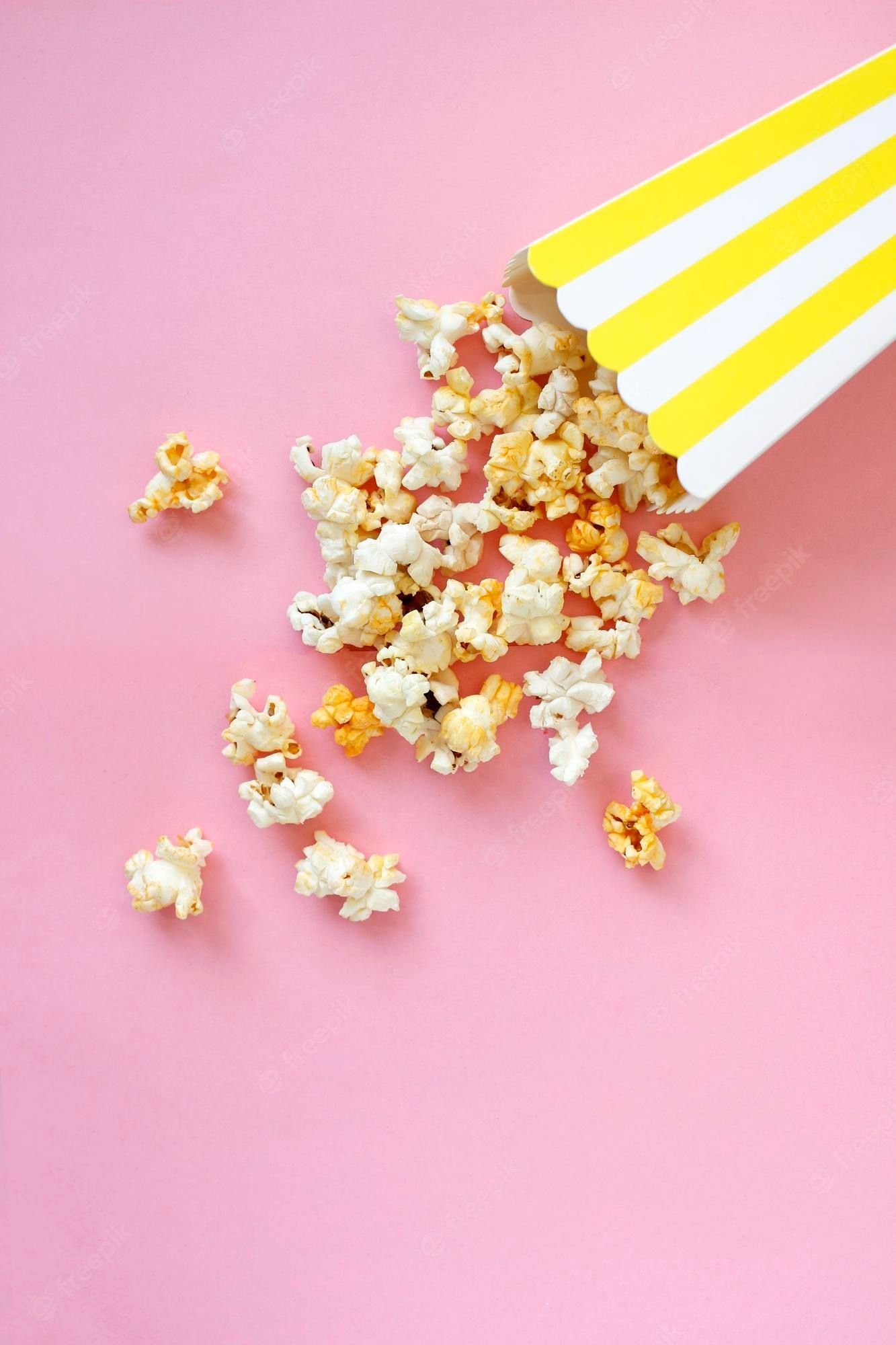 Popcorn spilling out of a yellow and white striped popcorn container on a pink background - Popcorn