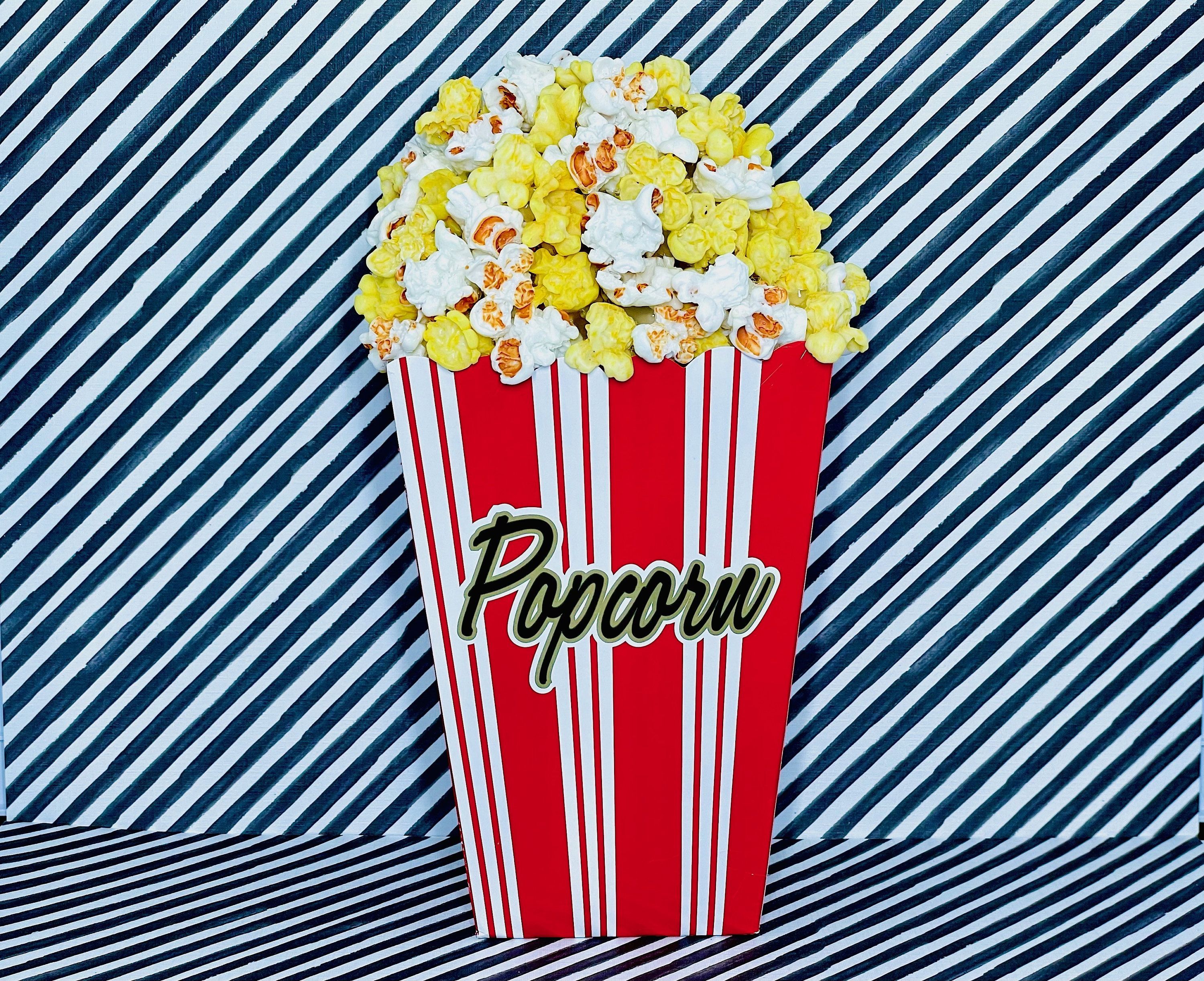 A large red and white popcorn box filled with popcorn on a blue and white striped background. - Popcorn
