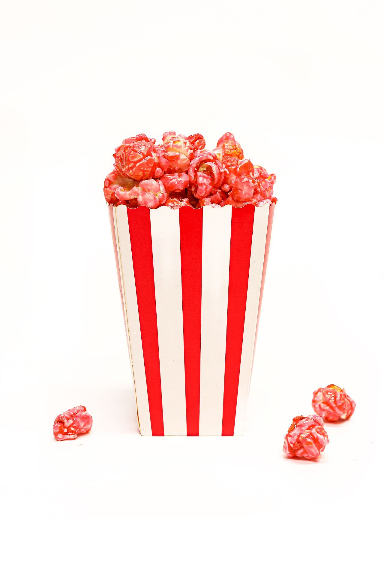 A red and white striped popcorn container with red popcorn spilling out of it. - Popcorn