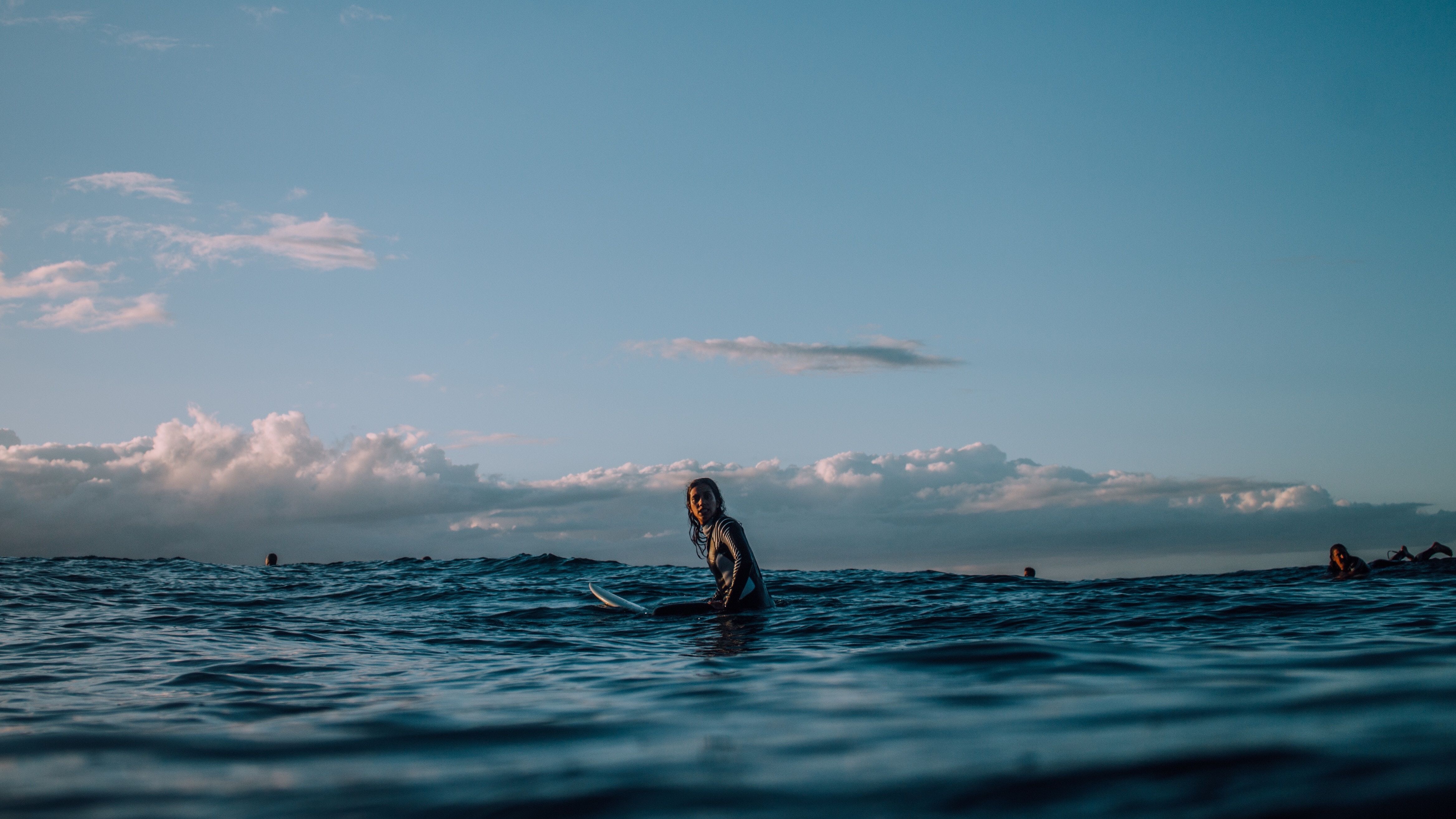 A woman sits on her surfboard in the ocean, surrounded by other surfers. The sky is blue and cloudy. - Surf